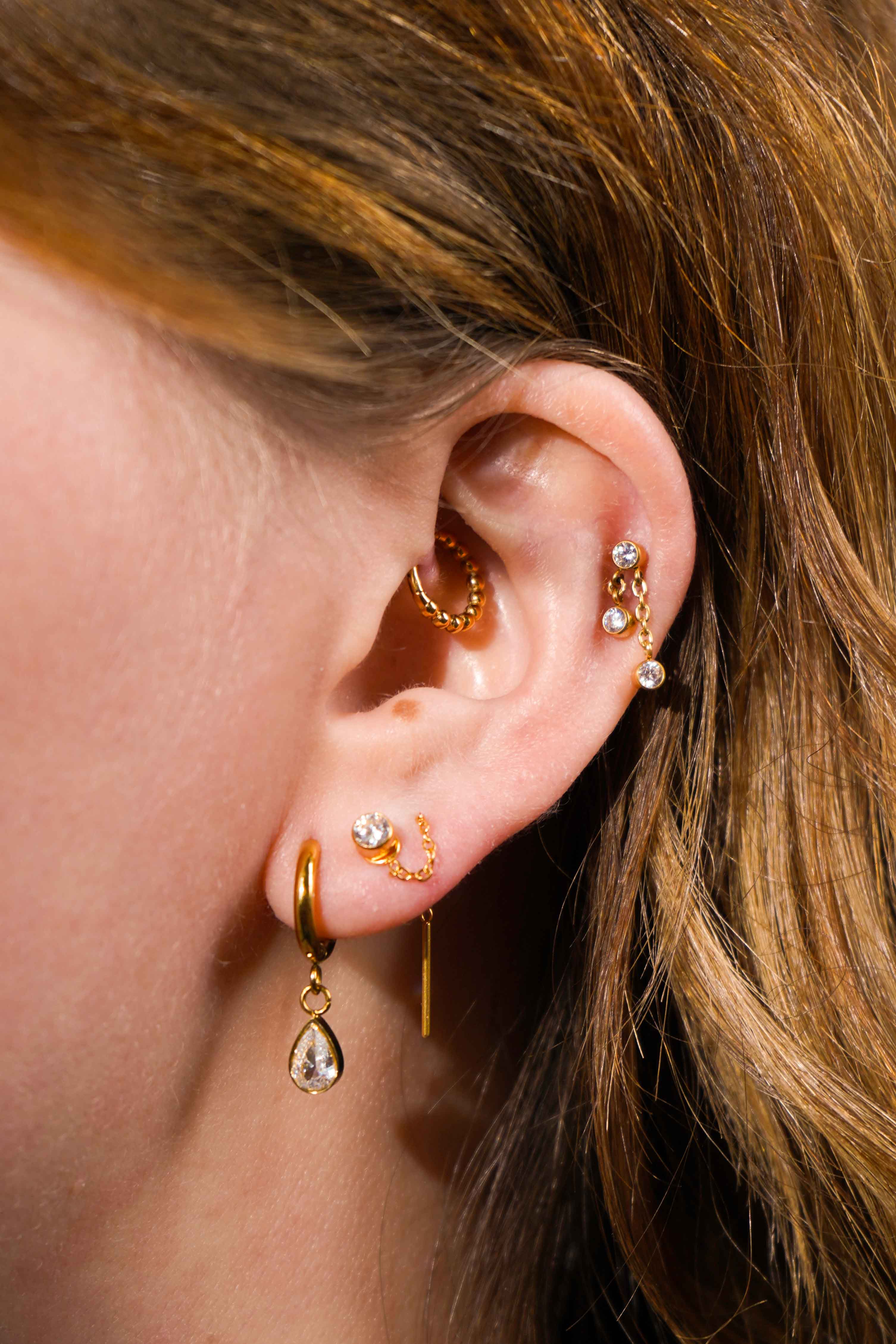 Close up of a model's ear, with multiple earrings including the featured product, a push-pin style flat back stud featuring a bezel-set round zirconia stone, clear in color. The flat back is made from ASTM F136 Implant Grade Titatnium, and is finished with 18k Gold PVD plating.