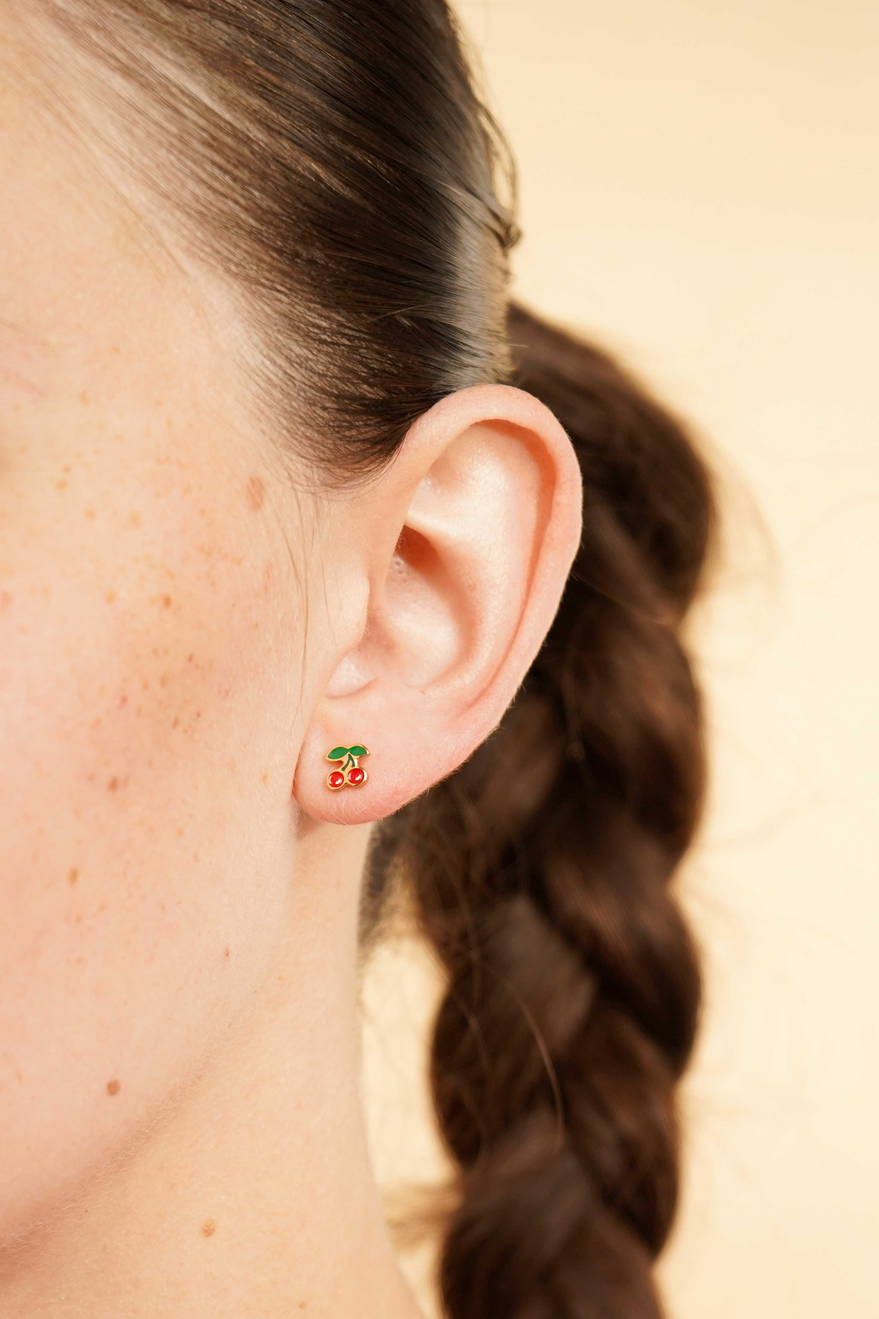 Close-up of an ear wearing a colorful enamel and gold double cherry flat back stud earring with a blurred background