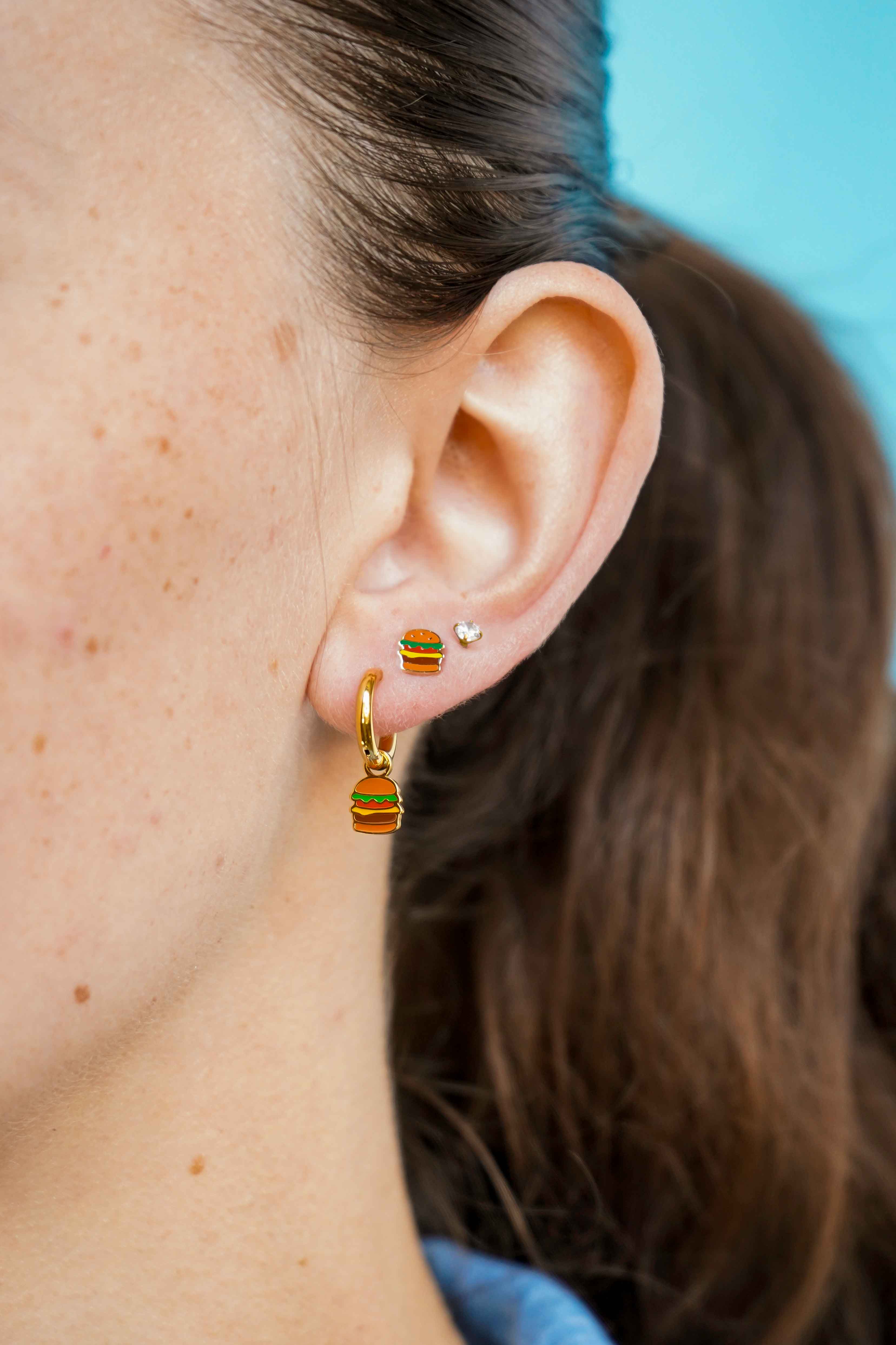 Close-up of an ear wearing a gold cheeseburger-shaped flat back stud earring and a gold hoop earring with a blue background