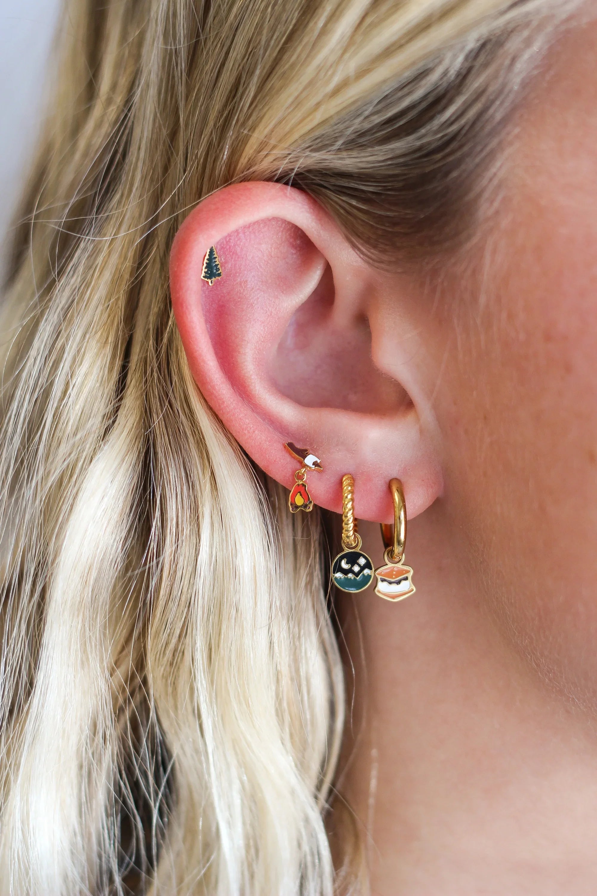 Close up of an ear with multiple piercings, featuring a green pine tree stud and a marshmallow on a stick over a dangled campfire stud, plus 2 gold hoops, 1 with a s'mores charm and the other with round night sky scene charm.