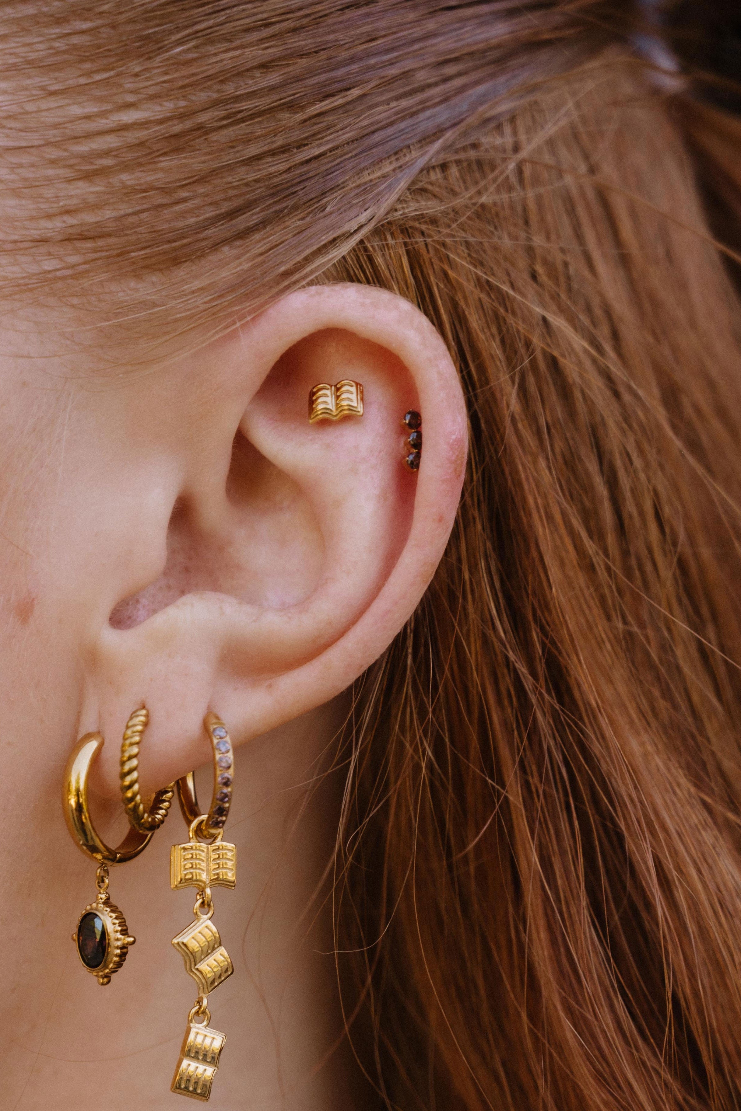 Close-up of a person's ear showcasing multiple gold piercings, including a small gold book charm earring with a dark stone in the lobe, a gold hoop, and a gold cartouche hoop with a small gold book charm.