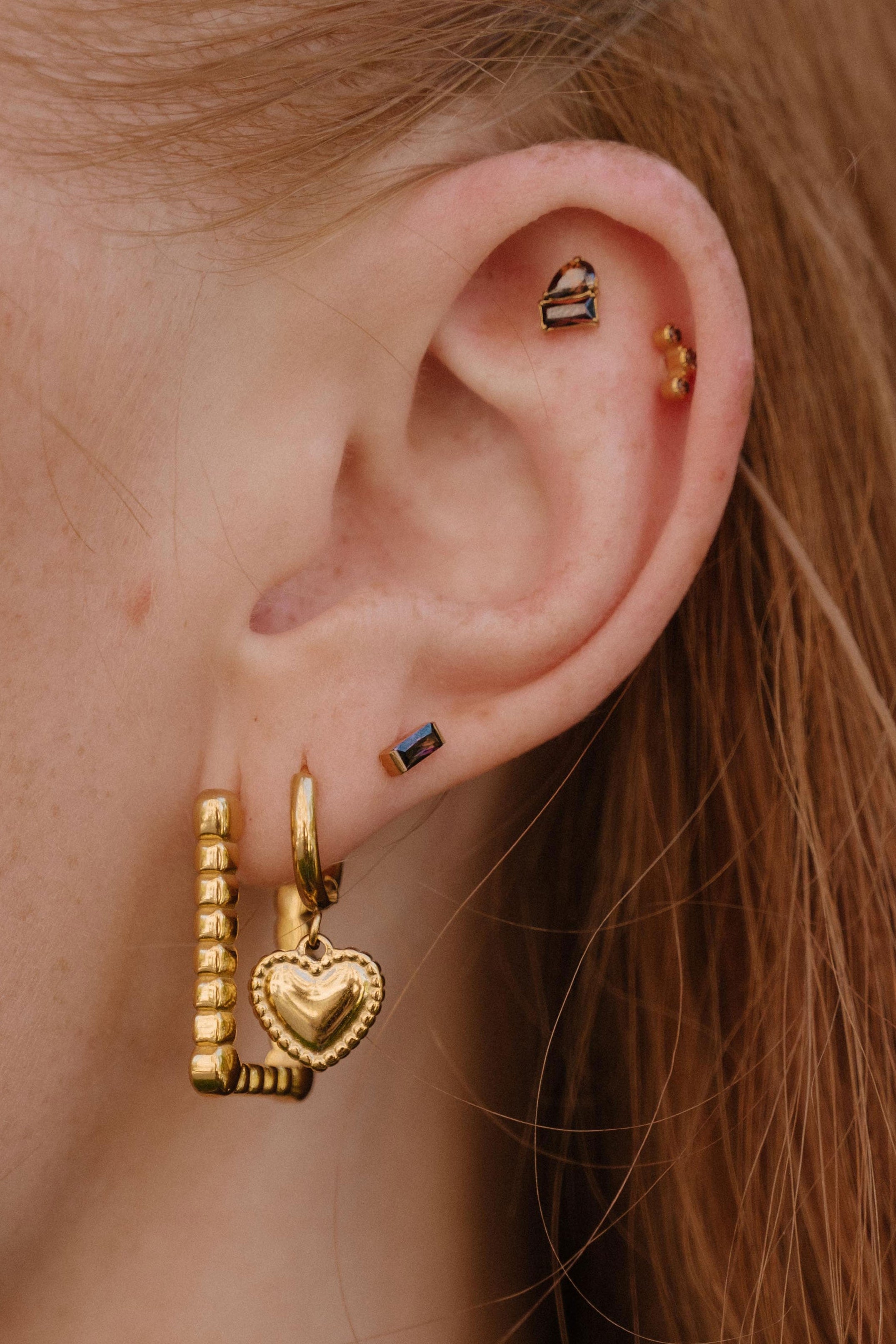 A close-up of a person's ear showcasing multiple gold piercings, including a large huggie hoop with a dangling beaded bar and heart charm, a small gold hoop, a rectangular dark blue stone stud, and ear cuffs.