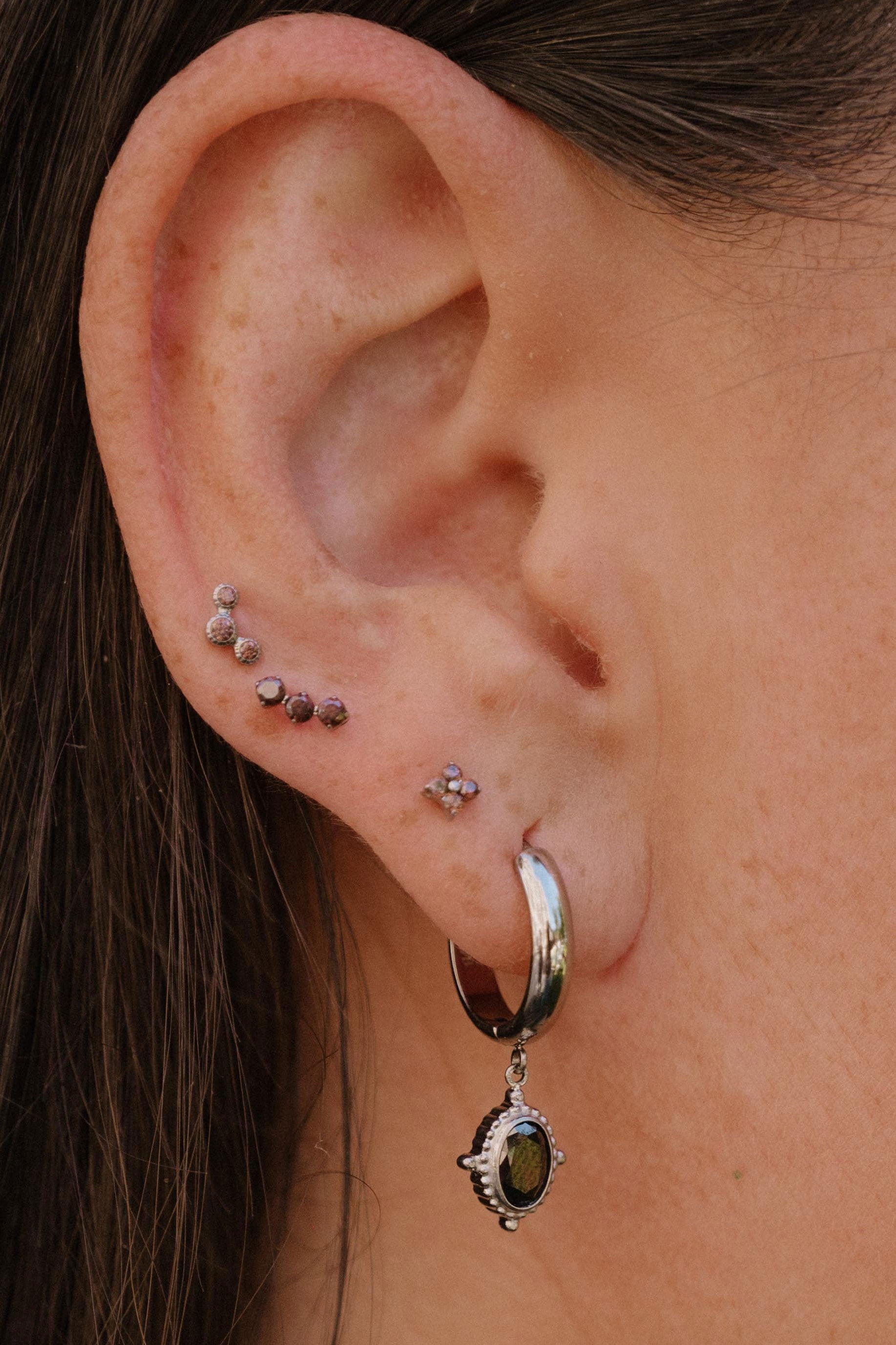 A close-up of a person's ear showcasing multiple piercings, including a silver hoop earring with a dangling brown stone and several small studs and piercings on the helix and lobe.