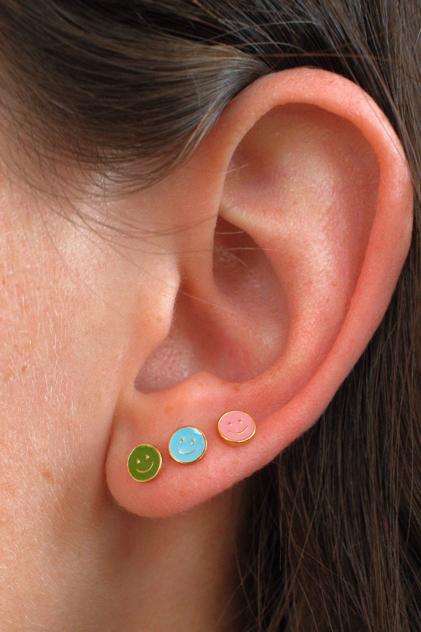 Close-up of a person's ear wearing three small smiley face stud earrings in green, light blue, and pink.