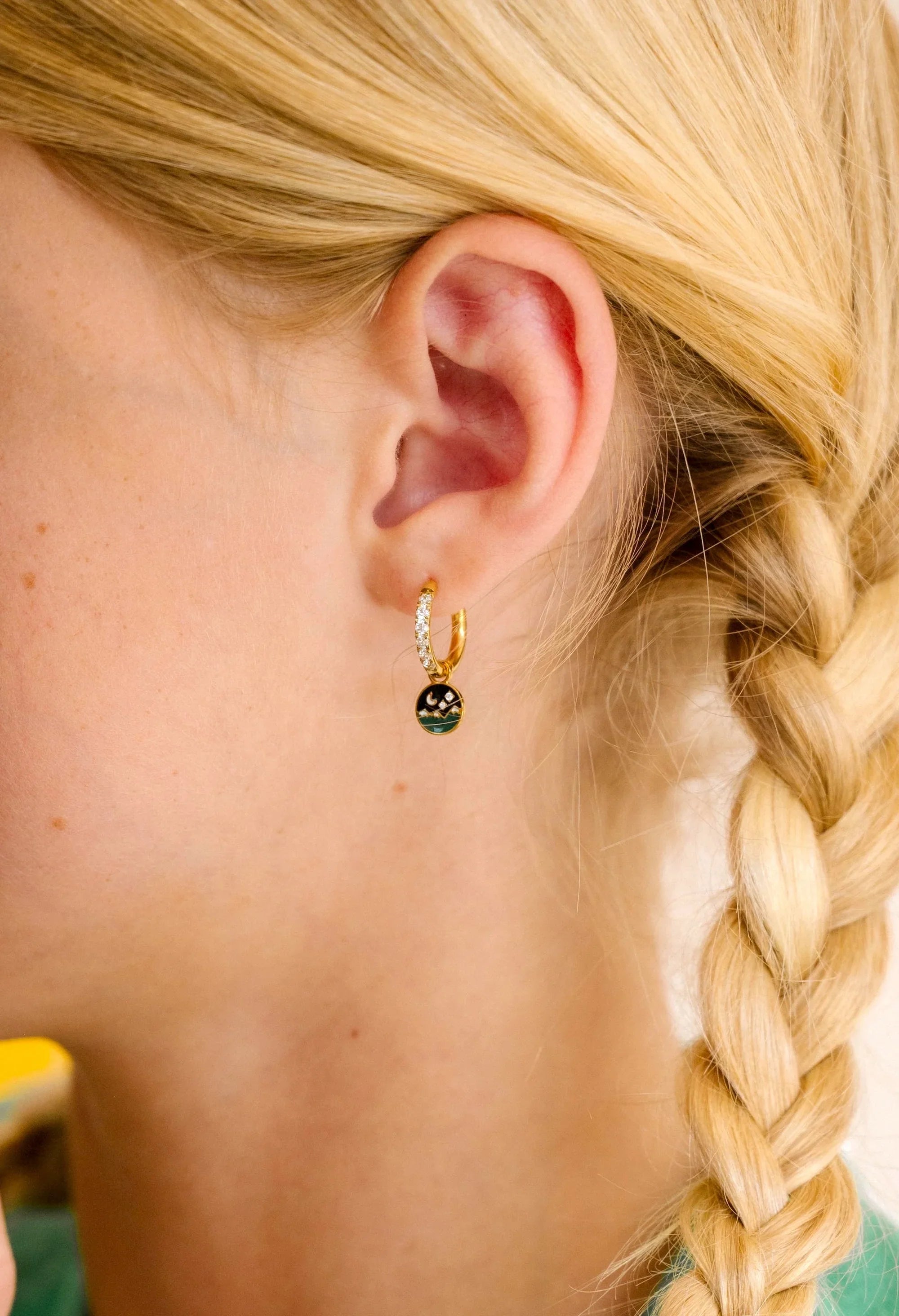 Close-up of an ear wearing a gold earring with a night sky scene made from enamel and clear gemstones, blonde hair in a braid.