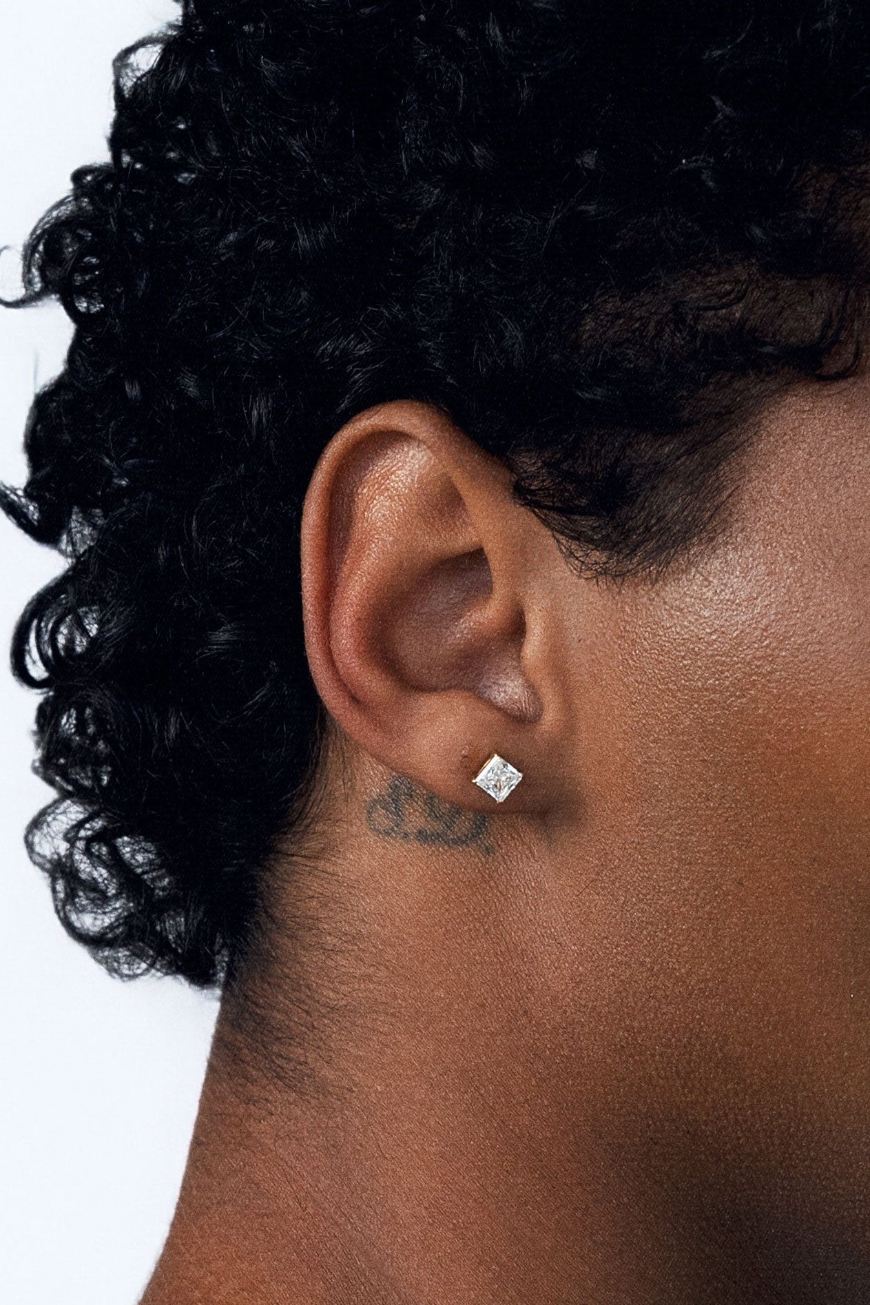 A close-up side profile of a person with curly black hair wearing a statement-sized, clear square-cut CZ stud earring on their earlobe, against a plain white background.