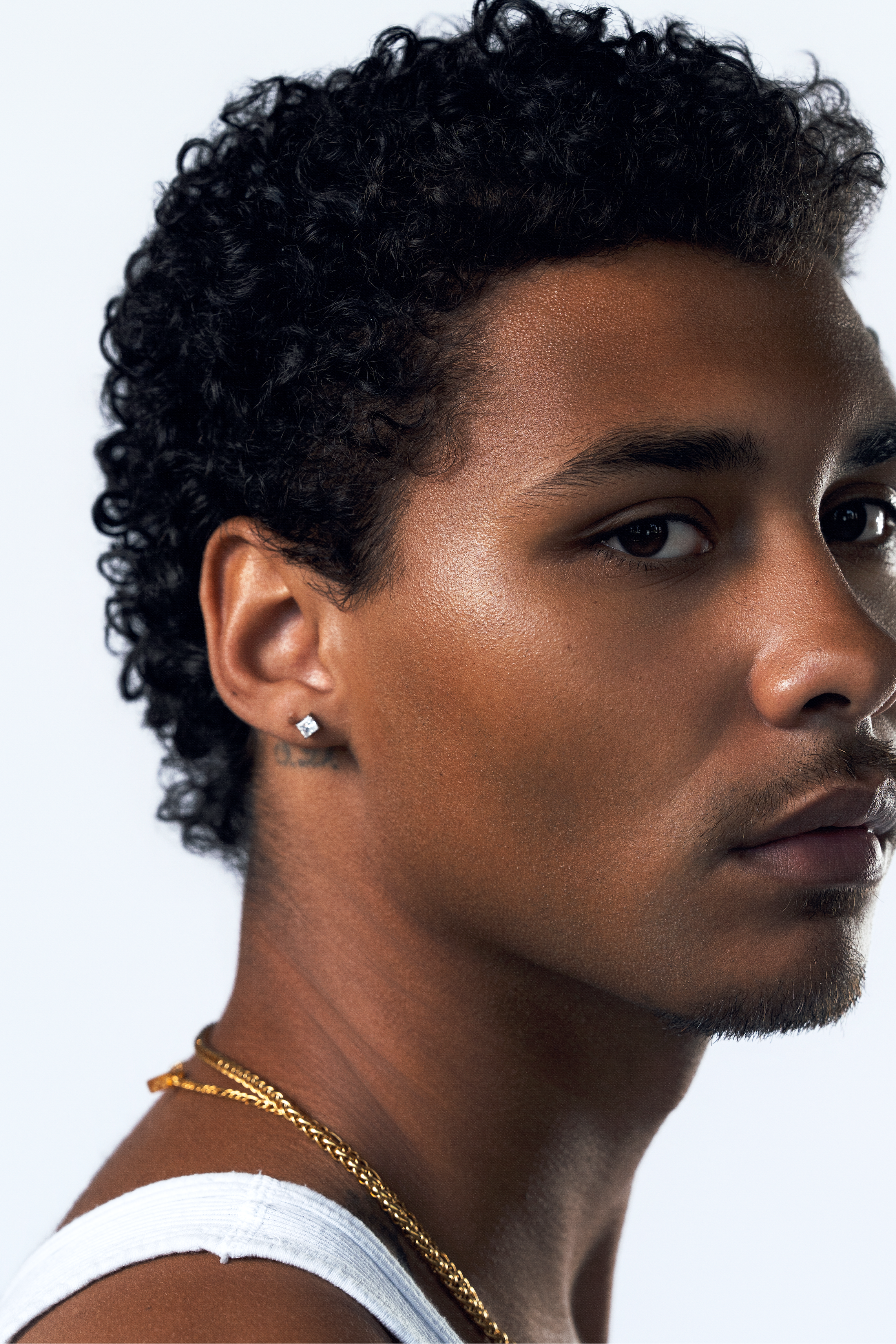 A close-up portrait of a man with curly black hair, wearing a white tank top, a gold chain necklace, and a small square cz stud earring, shown against a plain light gray background.