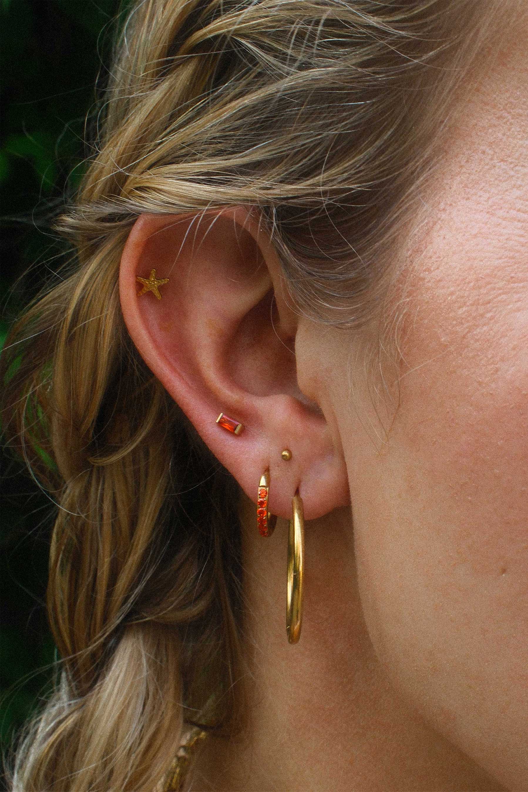 Close-up of an ear with gold hoop earrings and orange studs against a blurred background.