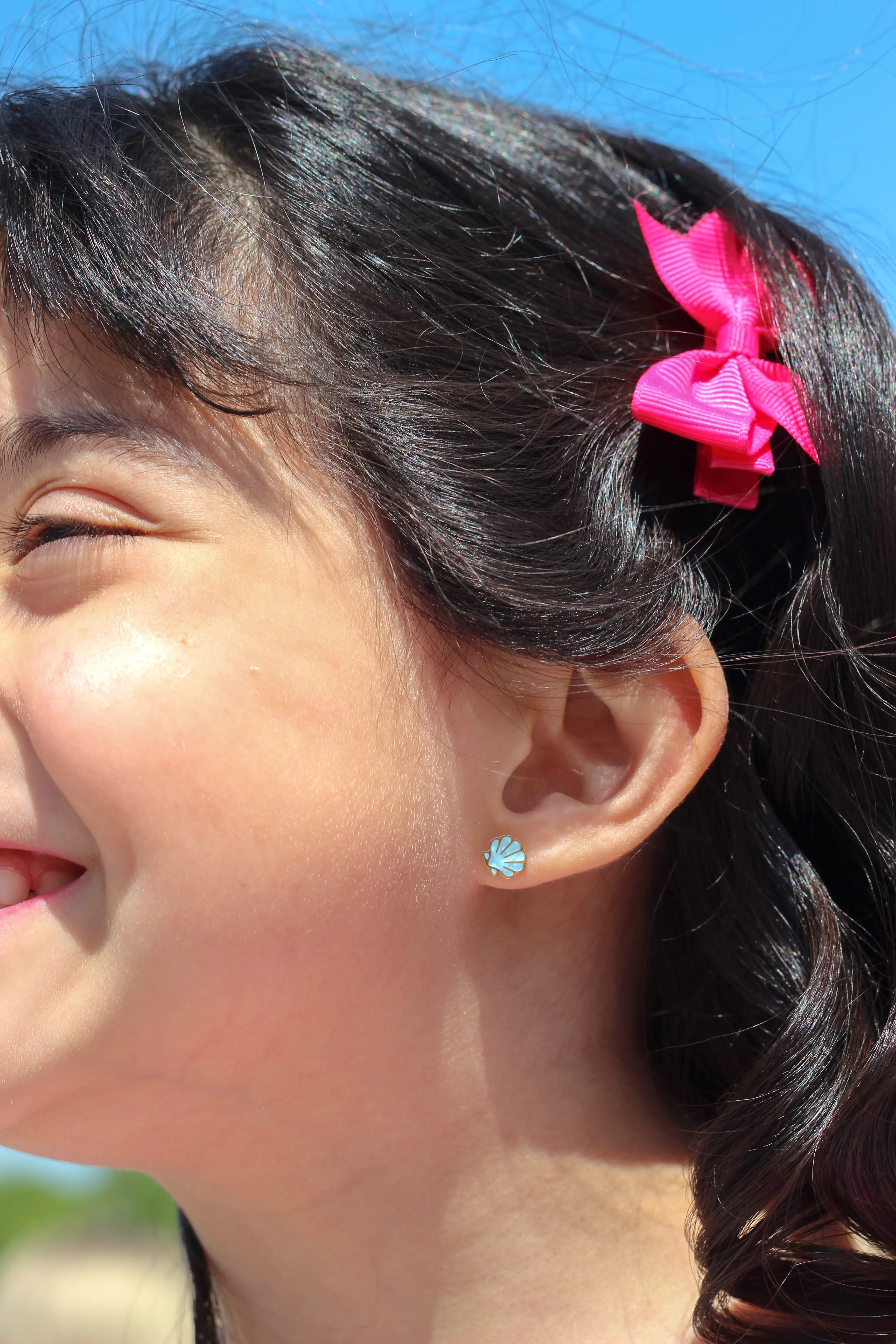 Close-up of a child with a pink hair clip and blue shell earring against a blue sky.