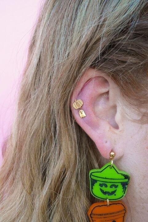 Close-up of an ear wearing a green pumpkin earring with a smiling face, against a pink background.