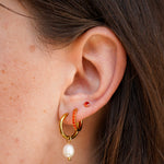 Close-up of a person's ear wearing a gold huggie hoop earring with a dangling white pearl and a small orange beaded accent, alongside a small red stud earring.