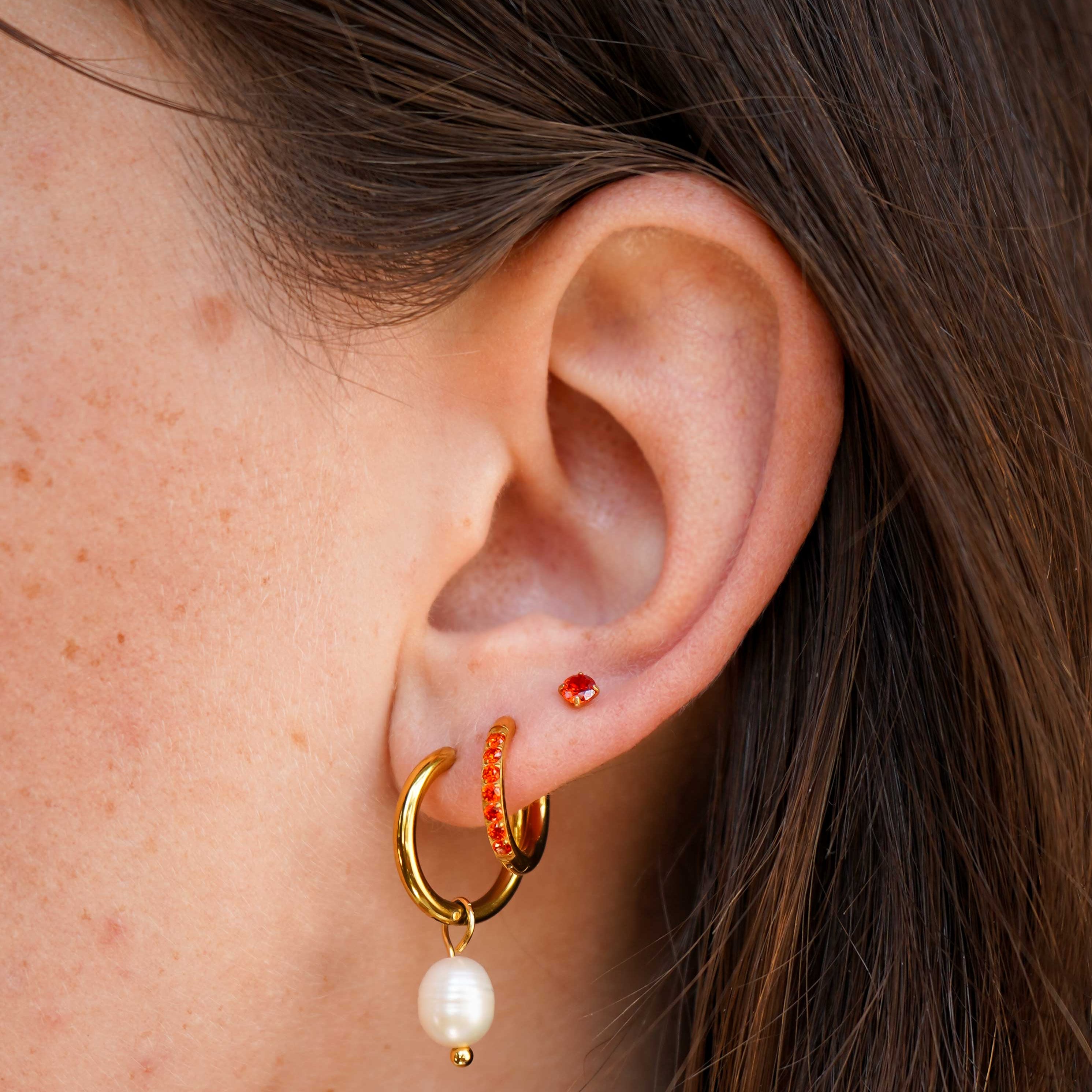 Close-up of a person's ear wearing a gold huggie hoop earring with a dangling white pearl and a small orange beaded accent, alongside a small red stud earring.