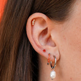 A close-up of a person's ear showcasing multiple earrings, including a silver huggie hoop with a dangling white pearl, a small orange gem stud, a silver and orange gem hoop, and a beaded cartilage flat back stud.