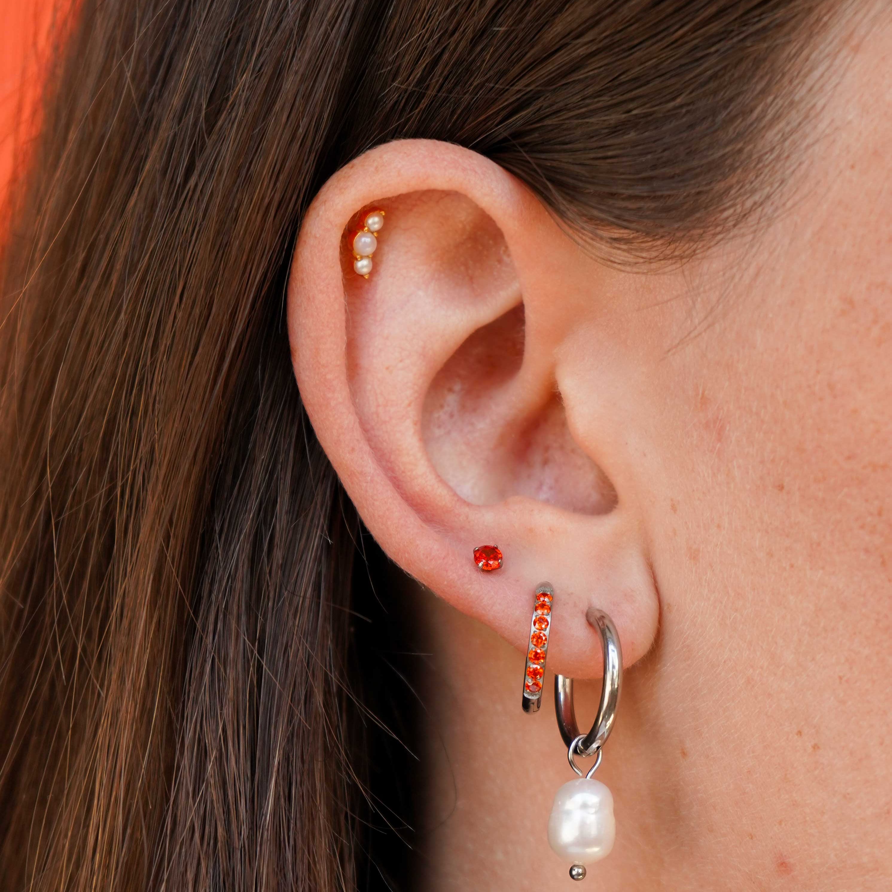 A close-up of a person's ear showcasing multiple earrings, including a silver huggie hoop with a dangling white pearl, a small orange gem stud, a silver and orange gem hoop, and a beaded cartilage flat back stud.