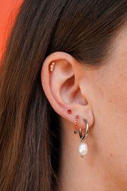 A close-up of a person's ear showcasing multiple earrings, including a silver huggie hoop with a dangling white pearl, a small orange gem stud, a silver and orange gem hoop, and a beaded cartilage flat back stud.