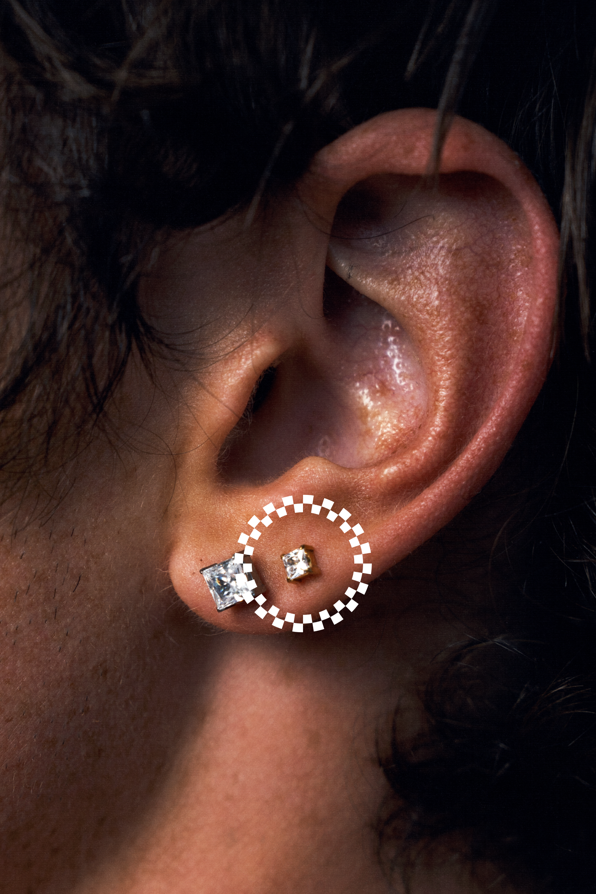 Close-up of a person's ear wearing two earrings: a square-cut CZ stud and a small square CZ stud, with a white dotted circle highlighting the smaller earring.