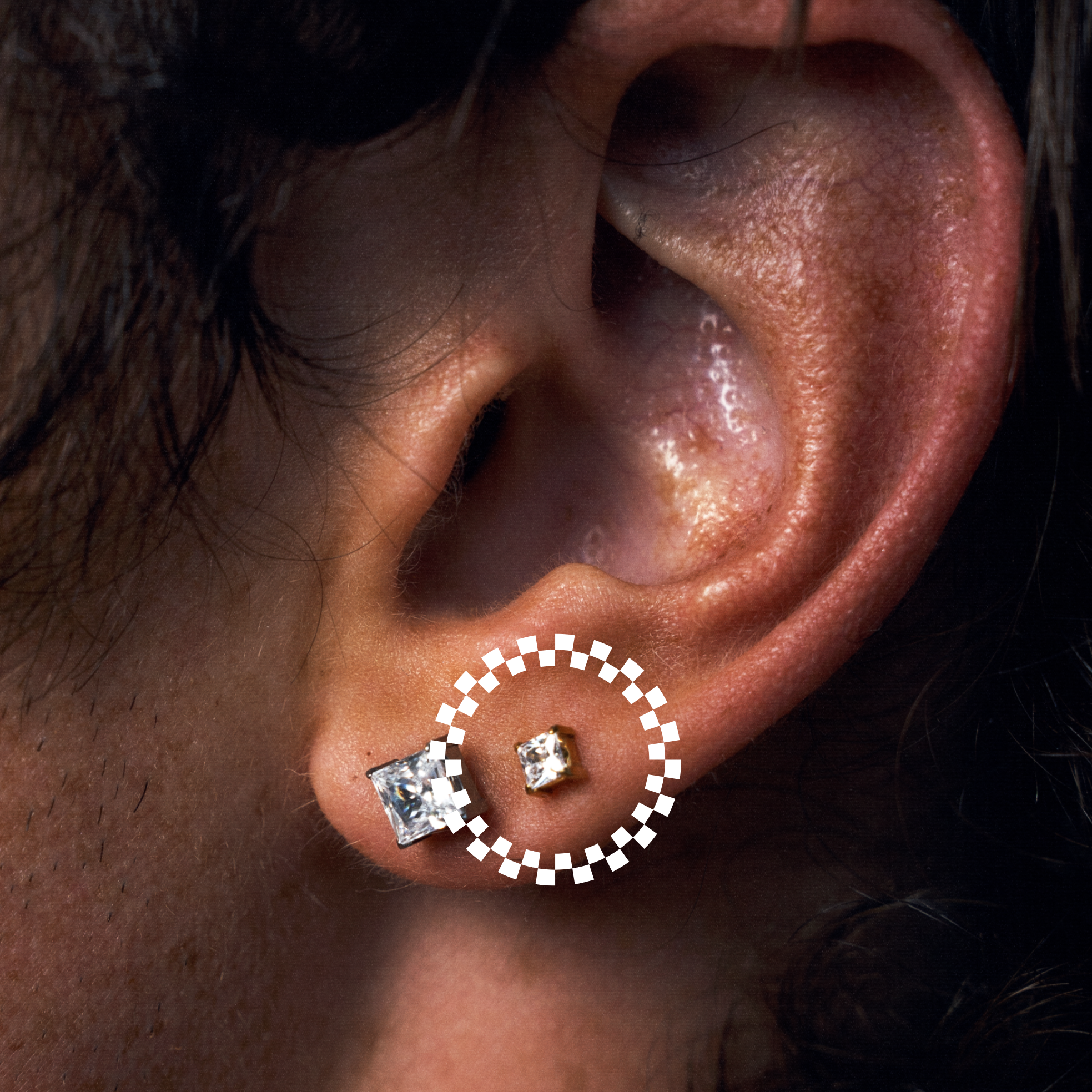 Close-up of a person's ear wearing two earrings: a square-cut CZ stud and a small square CZ stud, with a white dotted circle highlighting the smaller earring.