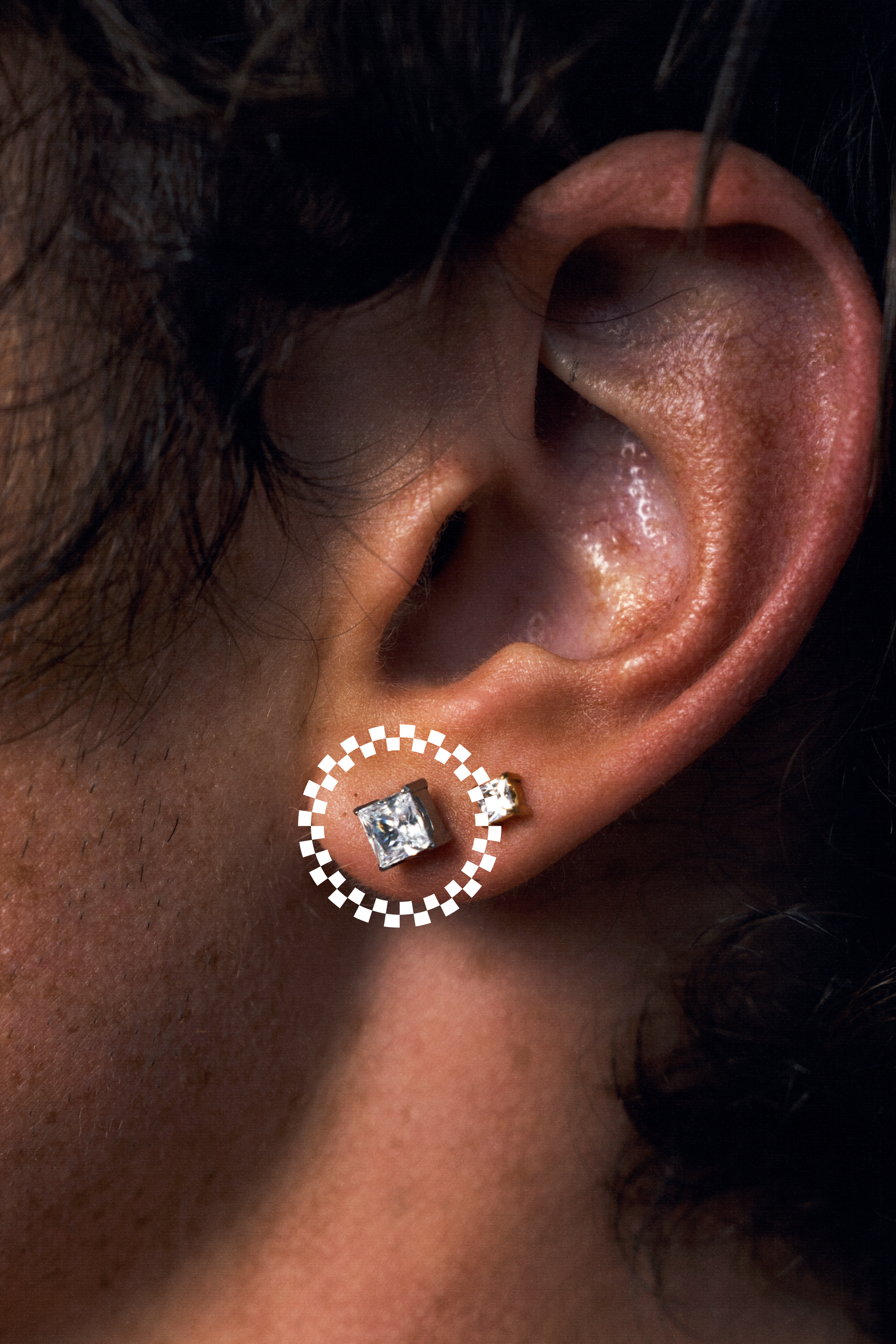 A close-up of a person's ear wearing a medium square stud earring with a clear gemstone and a small matching stud behind it. A dotted white circle highlights the front earring.