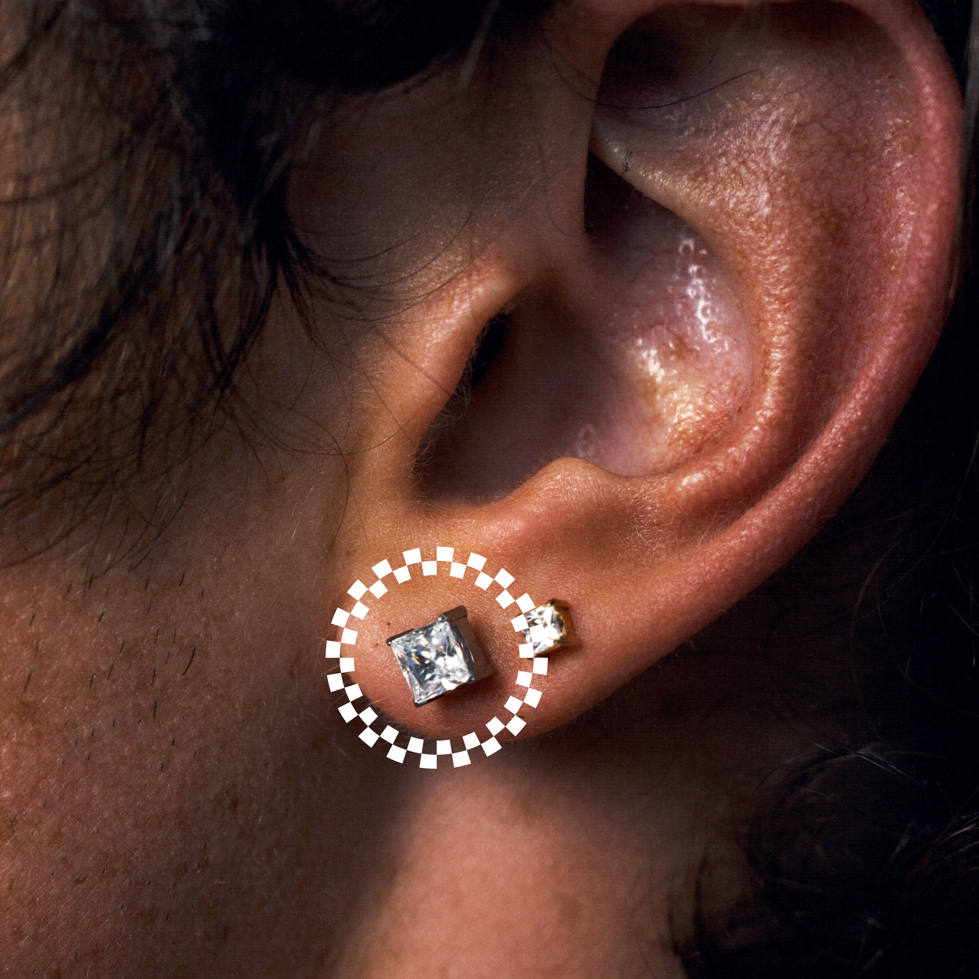 A close-up of a person's ear wearing a medium square stud earring with a clear gemstone and a small matching stud behind it. A dotted white circle highlights the front earring.