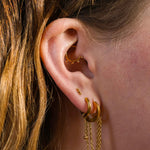 Close up of a model's ear with multiple earrings, including an earring charm featuring a length of metal chain with a metal ball dangling at the bottom. The charm is made from ASTM F136 Implant Grade titanium, and finished with 18K Gold PVD Plating