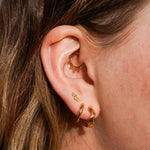 Close up of a model wearing multiple earrings, including clicker-style hoop featuring a series of round metal details that radiate from the main hoop in the Daith piercing. Made from ASTM F136 Implant-Grade Titanium, finished with 18K Gold PVD plating