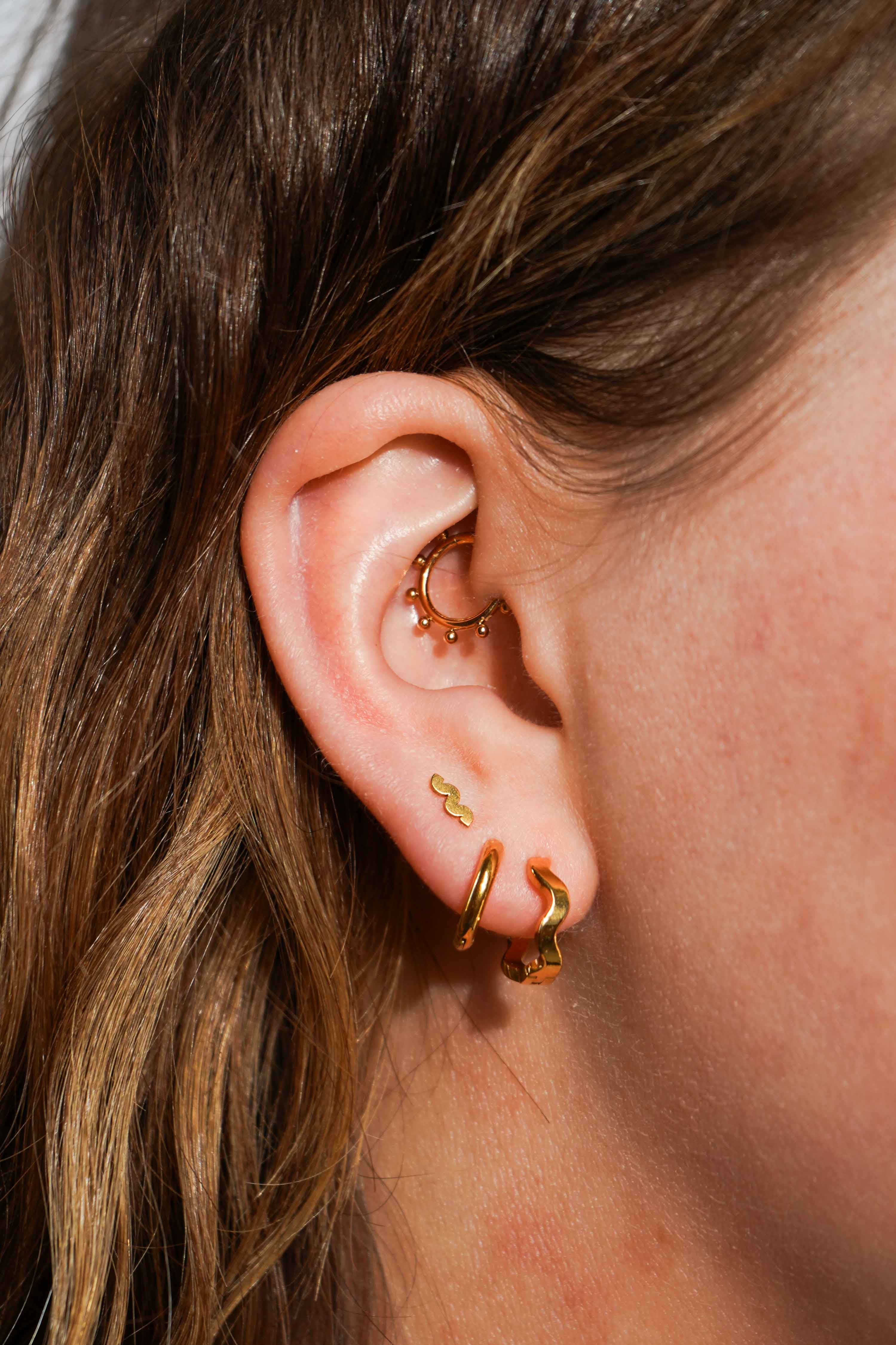 Close up of a model wearing multiple earrings, including clicker-style hoop featuring a series of round metal details that radiate from the main hoop in the Daith piercing. Made from ASTM F136 Implant-Grade Titanium, finished with 18K Gold PVD plating