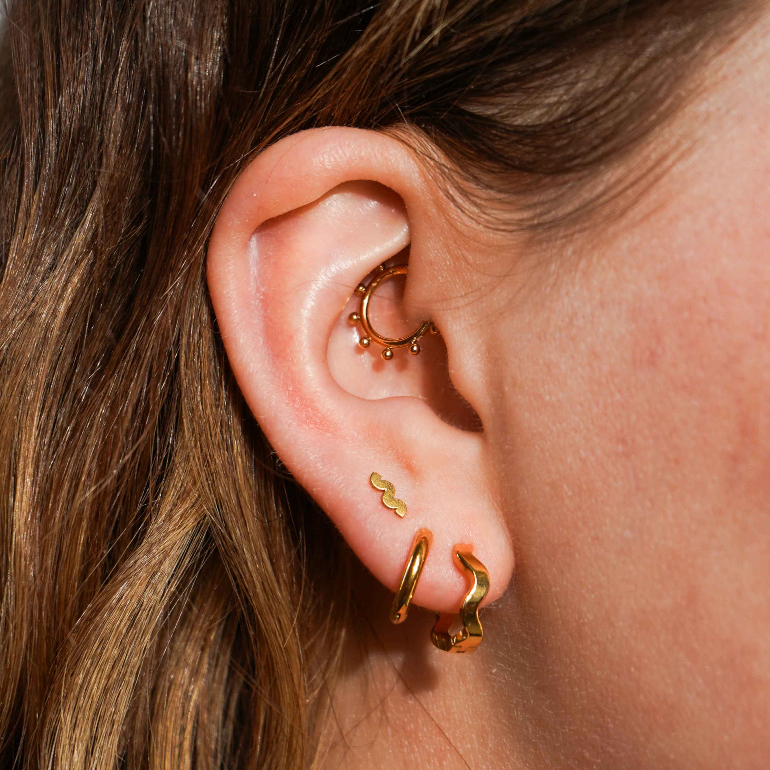 A close up of a models ear with multiple earrings, including a pair of hoop earrings that feature a wavy, rippled design. Hoops are made from ASTM F136 Implant Grade Titanium and are finished with 18k Gold PVD plating.