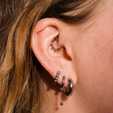 Close up of a model wearing multiple earrings, including clicker-style hoop featuring a series of round metal details that radiate from the main hoop in the Daith piercing. Made from ASTM F136 Implant-Grade Titanium