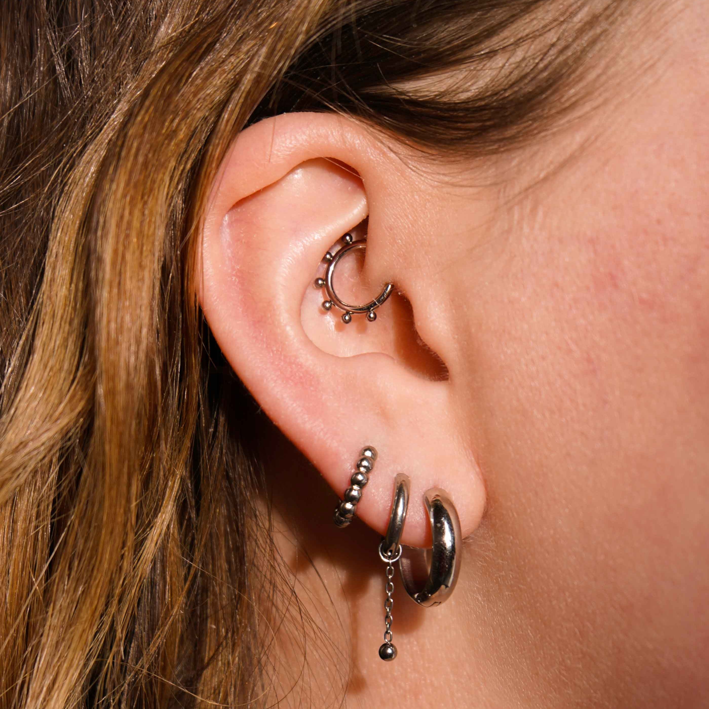 Close up of a model's ear with multiple earrings, including an earring charm featuring a length of metal chain with a metal ball dangling at the bottom. The charm is made from ASTM F136 Implant Grade titanium.