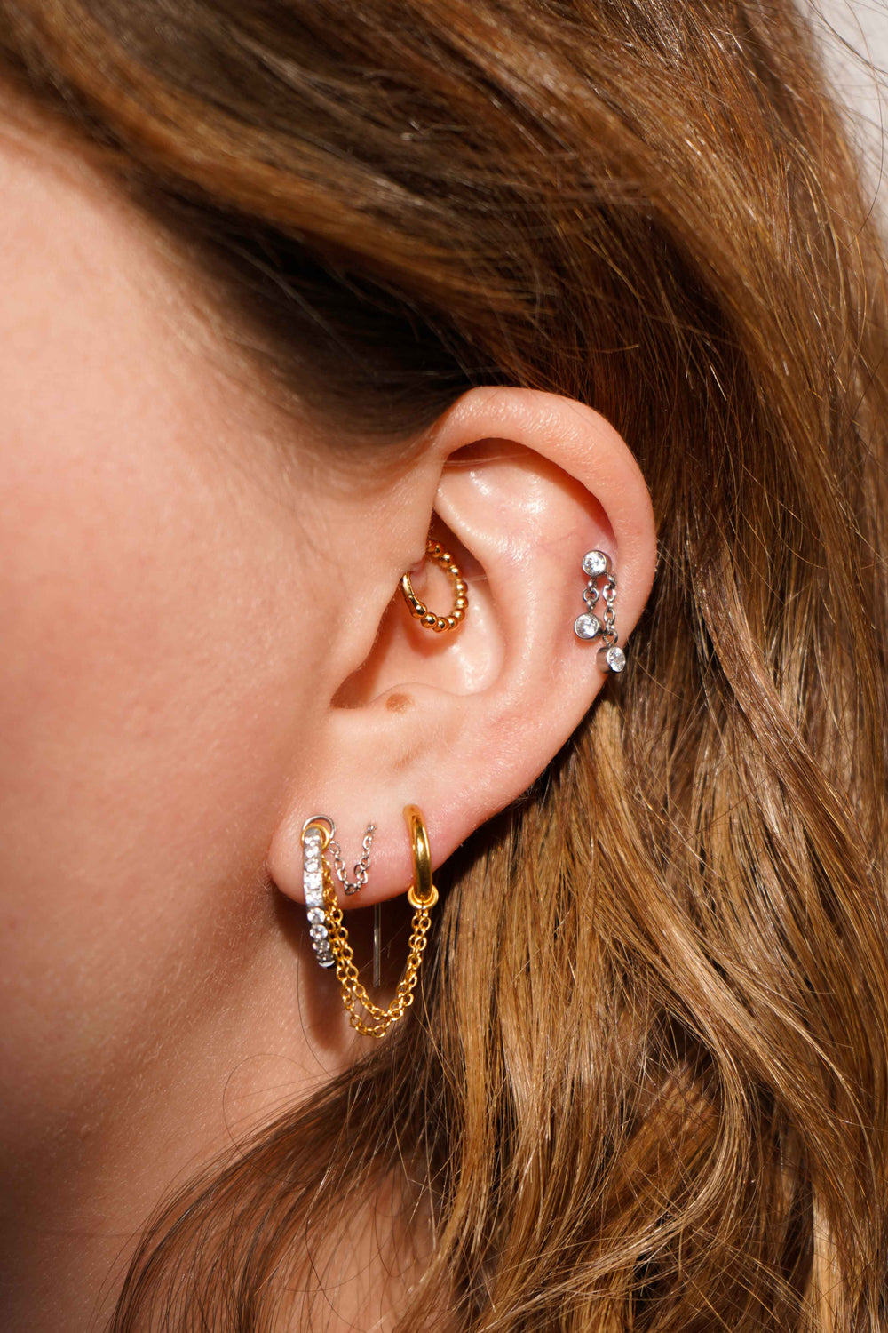 Close up of a model's ear with multiple earrings, including an earring accessory that features two layered chains attached to 2 small rings, and linked to hang between a hoop and flat back stud. Made from 316L Surgical Steel and finished with 18K Gold PVD plating.