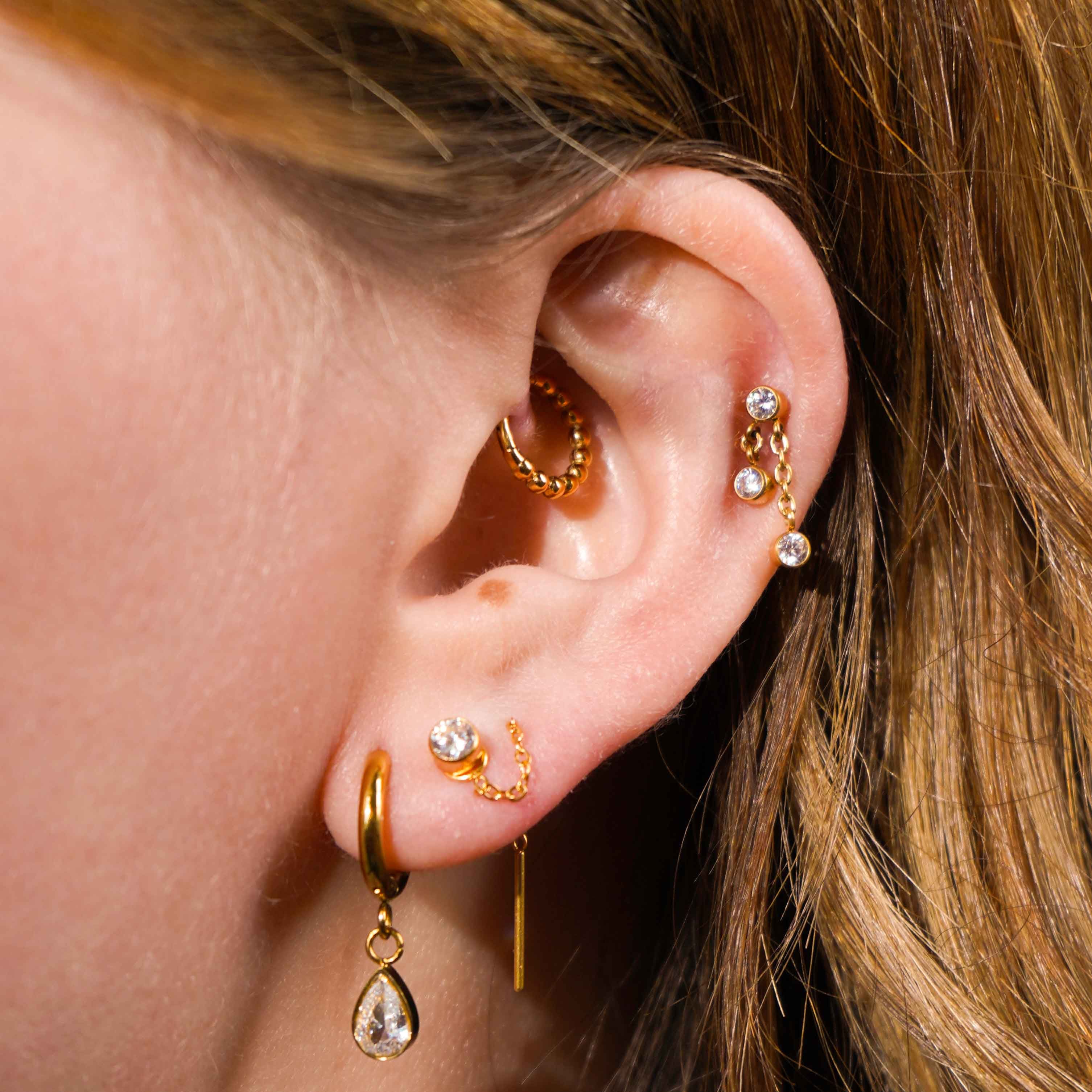 Close up of a model's ear, with multiple earrings including the featured product, a push-pin style flat back stud featuring a bezel-set round zirconia stone, clear in color, with 2 additional stones of the same style and color that hang from chains attached to the main stone. The flat back is made from ASTM F136 Implant Grade Titatnium, and is finished with 18k Gold PVD plating. 