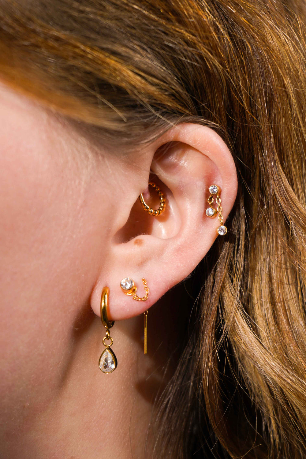 Close up of a model's ear, with multiple earrings including the featured product, a push-pin style flat back stud featuring a bezel-set round zirconia stone, clear in color, with 2 additional stones of the same style and color that hang from chains attached to the main stone. The flat back is made from ASTM F136 Implant Grade Titatnium, and is finished with 18k Gold PVD plating. 
