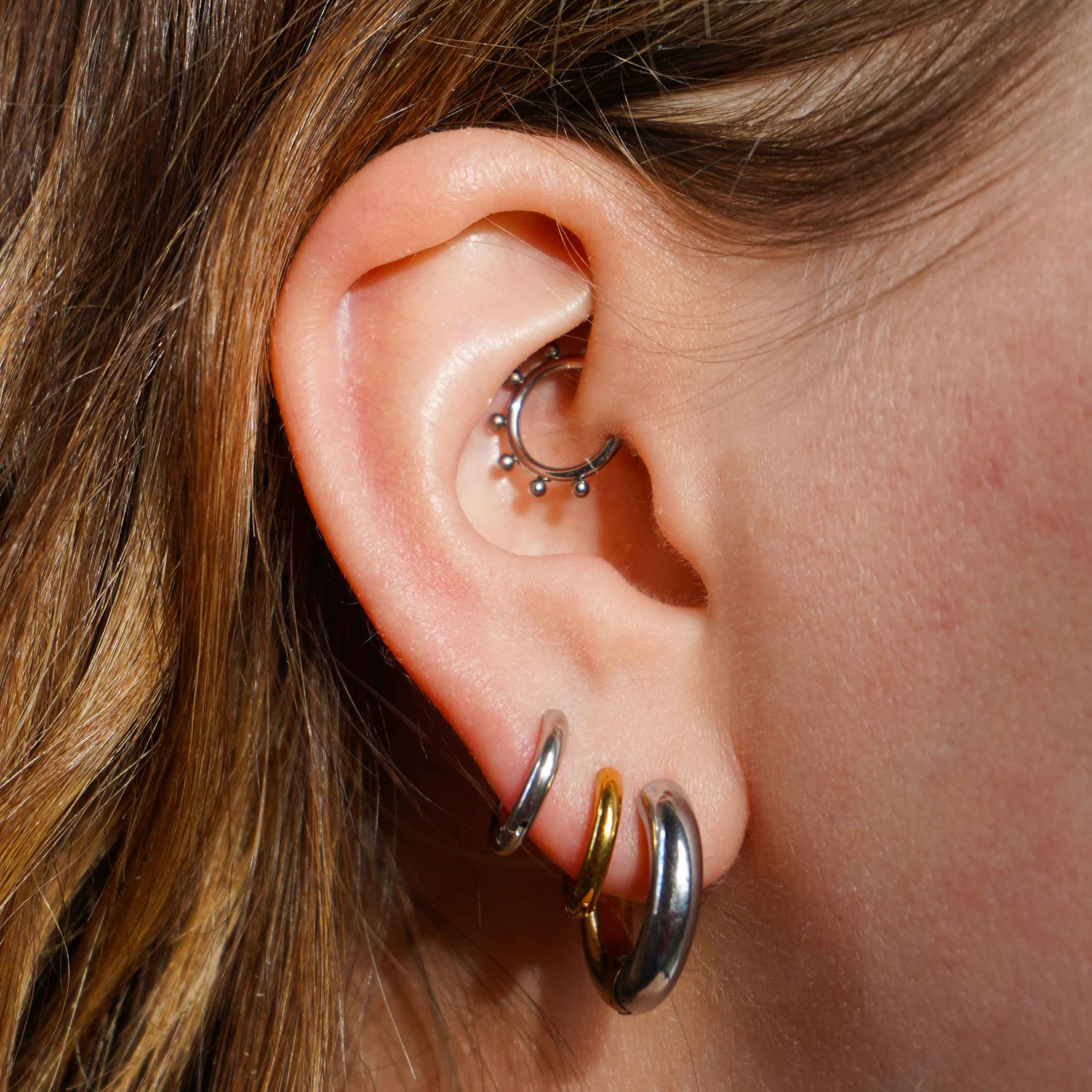 Close up of a model wearing multiple earrings, including clicker-style hoop featuring a series of round metal details that radiate from the main hoop in the Daith piercing. Made from ASTM F136 Implant-Grade Titanium