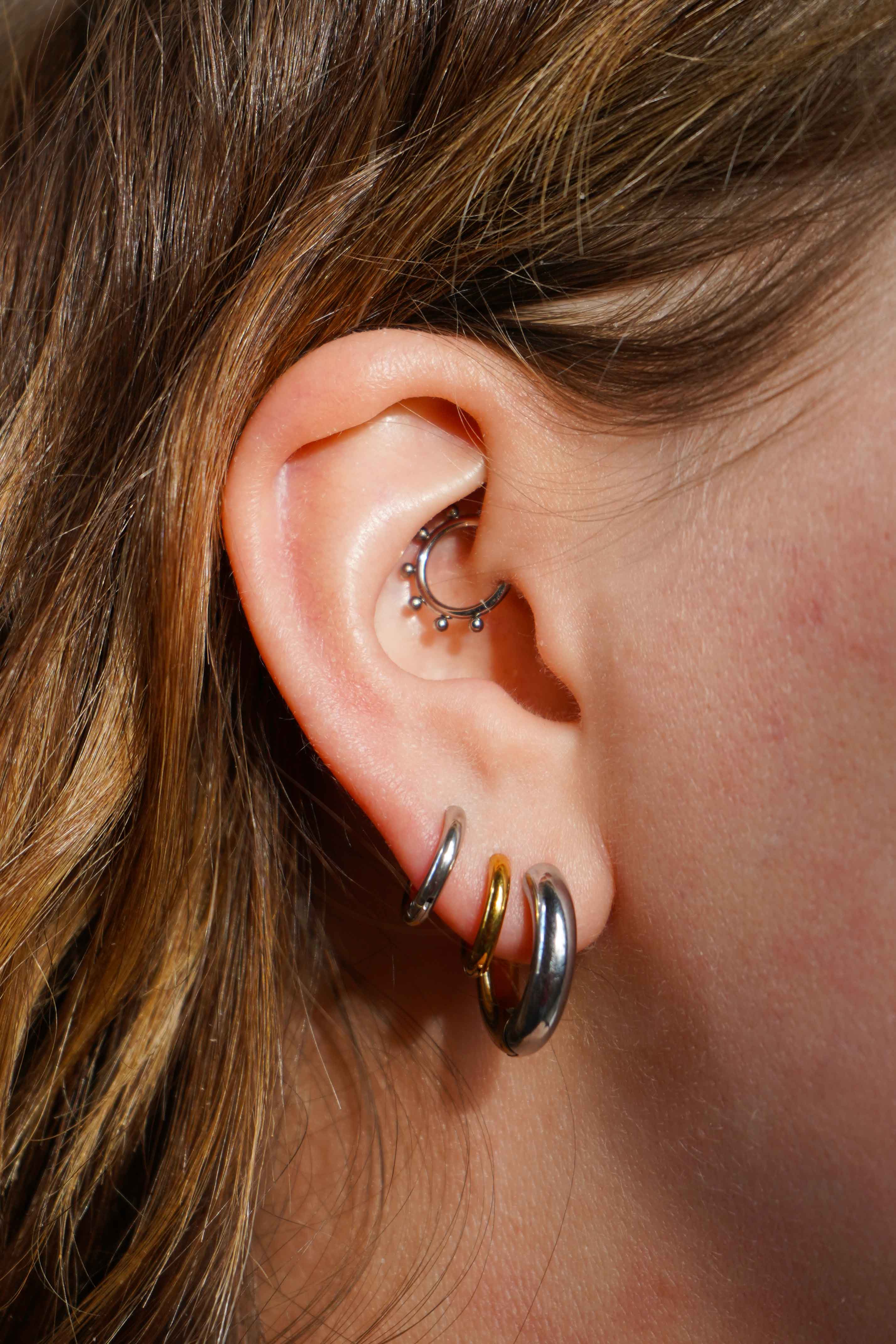 Close up of a model wearing multiple earrings, including clicker-style hoop featuring a series of round metal details that radiate from the main hoop in the Daith piercing. Made from ASTM F136 Implant-Grade Titanium