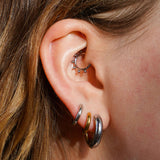 A close up of a model's ear wearing multiple earrings, including a hoop earring that features a split design where one half of the hoop is silver in color and the other is gold. The hoop is made from ASTM F136 Implant Grade Titanium and the gold half is finished with 18k Gold PVD plating.
