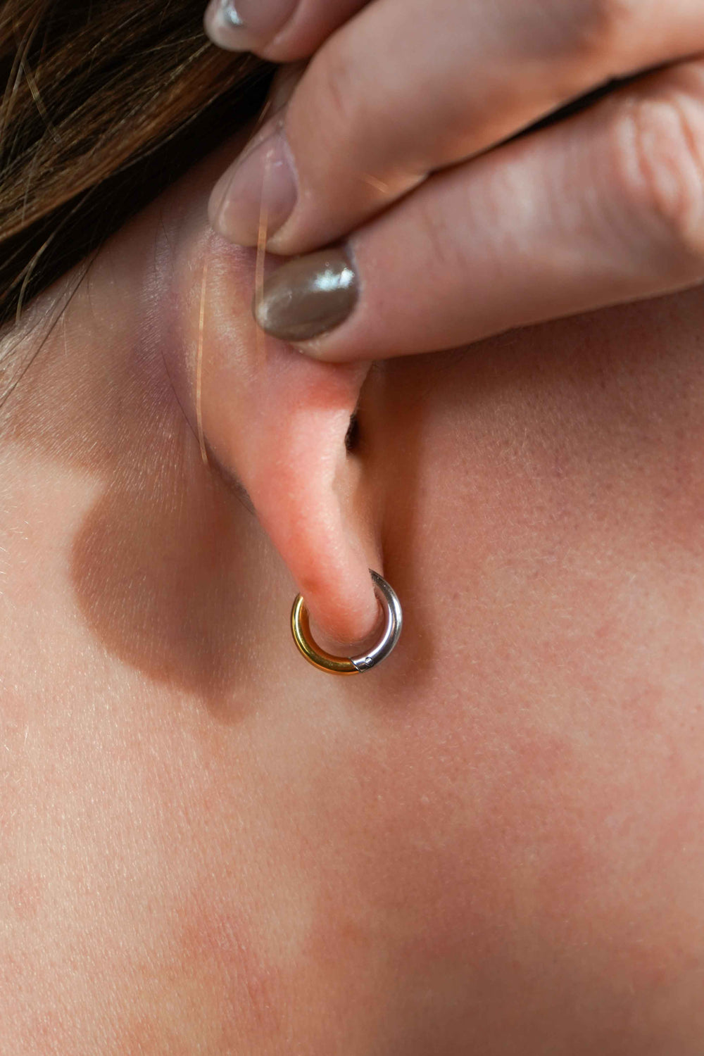 A close up of a model's ear wearing a hoop earring that features a split design where one half of the hoop is silver in color and the other is gold. Hoops are made from ASTM F136 Implant Grade Titanium and the gold half is finished with 18k Gold PVD plating.