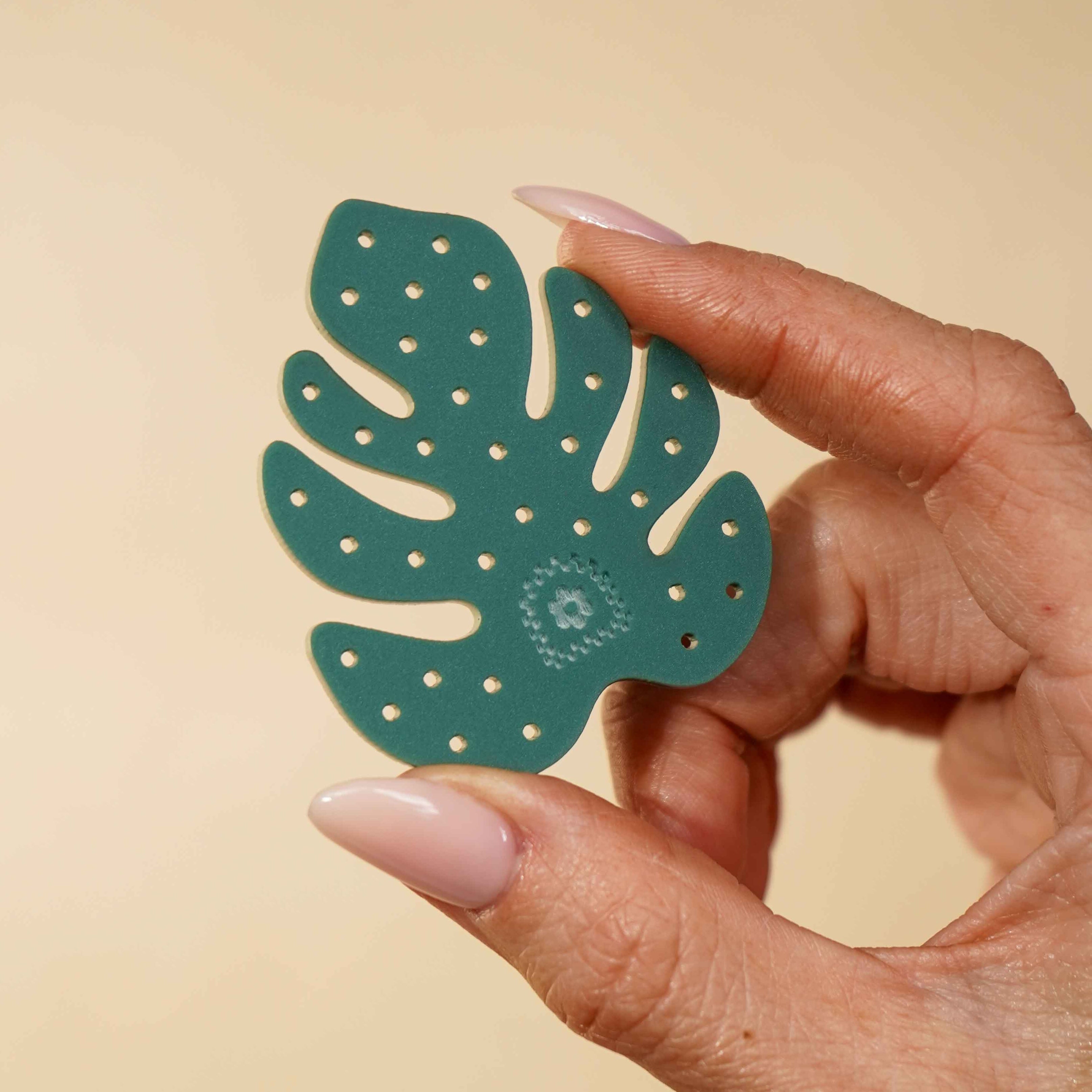 Hand holding a green leaf-shaped acrylic piece with little circle cutouts on a beige background