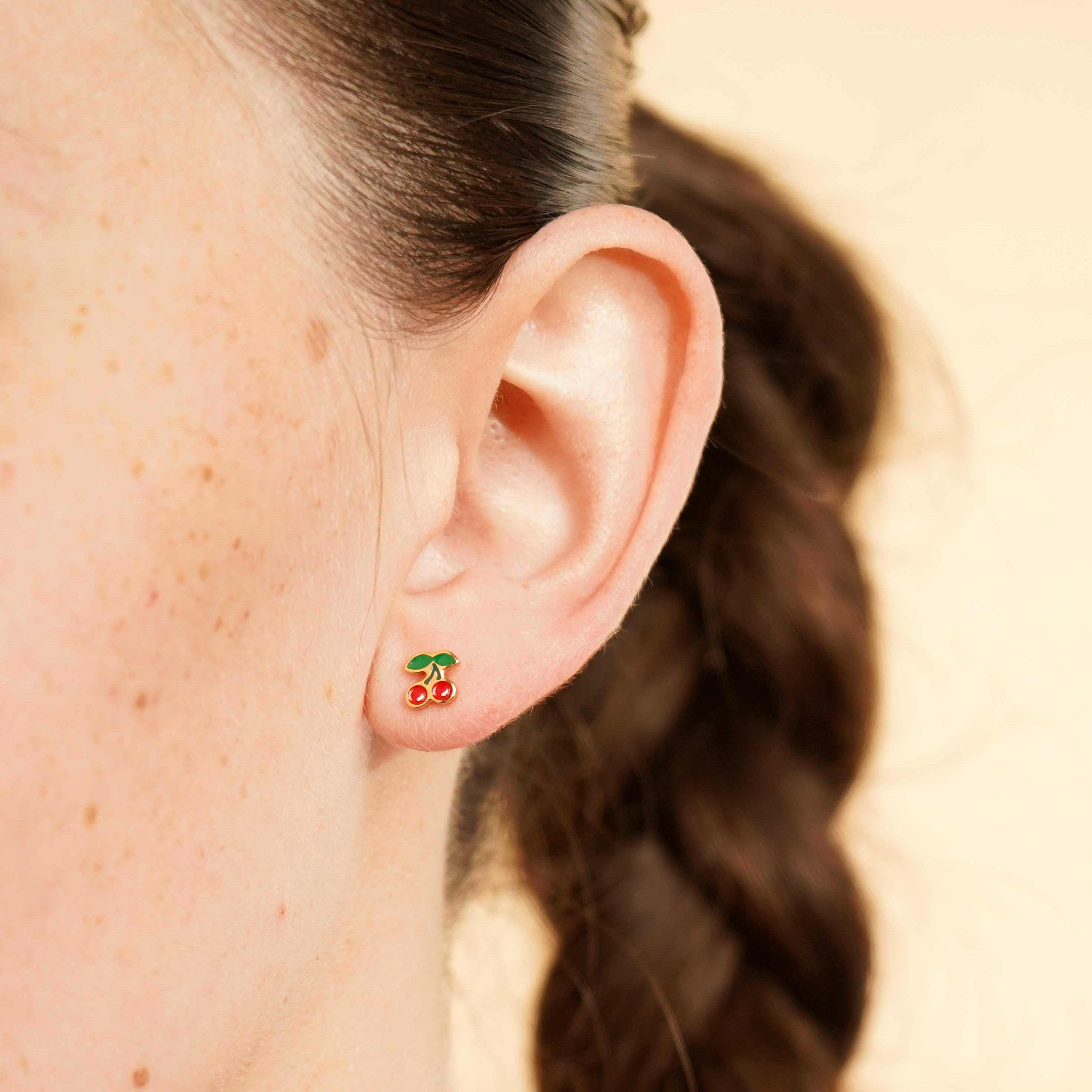 Close-up of an ear wearing a colorful enamel and gold double cherry flat back stud earring with a blurred background
