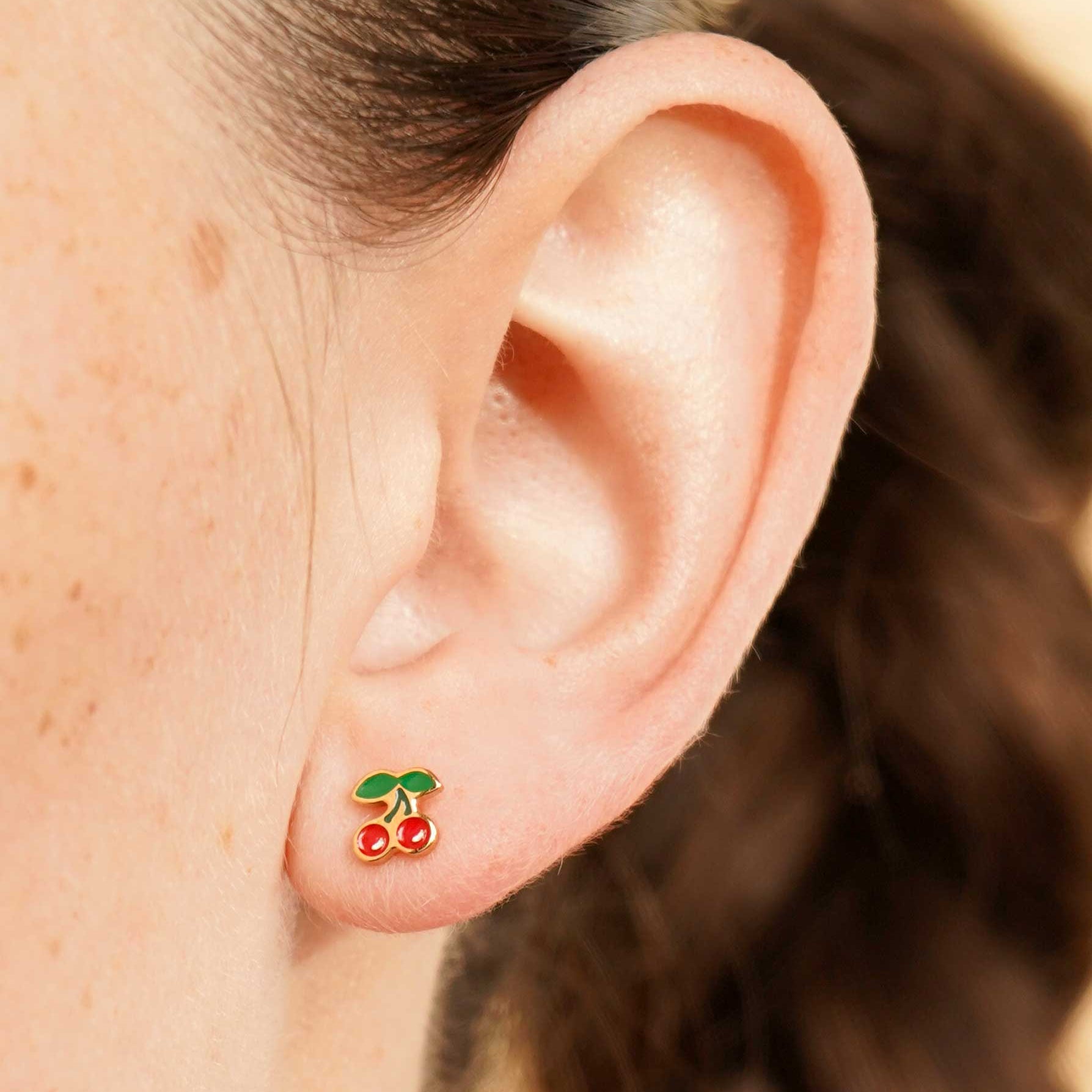 Close-up of an ear wearing a colorful enamel and gold double cherry flat back stud earring with a blurred background
