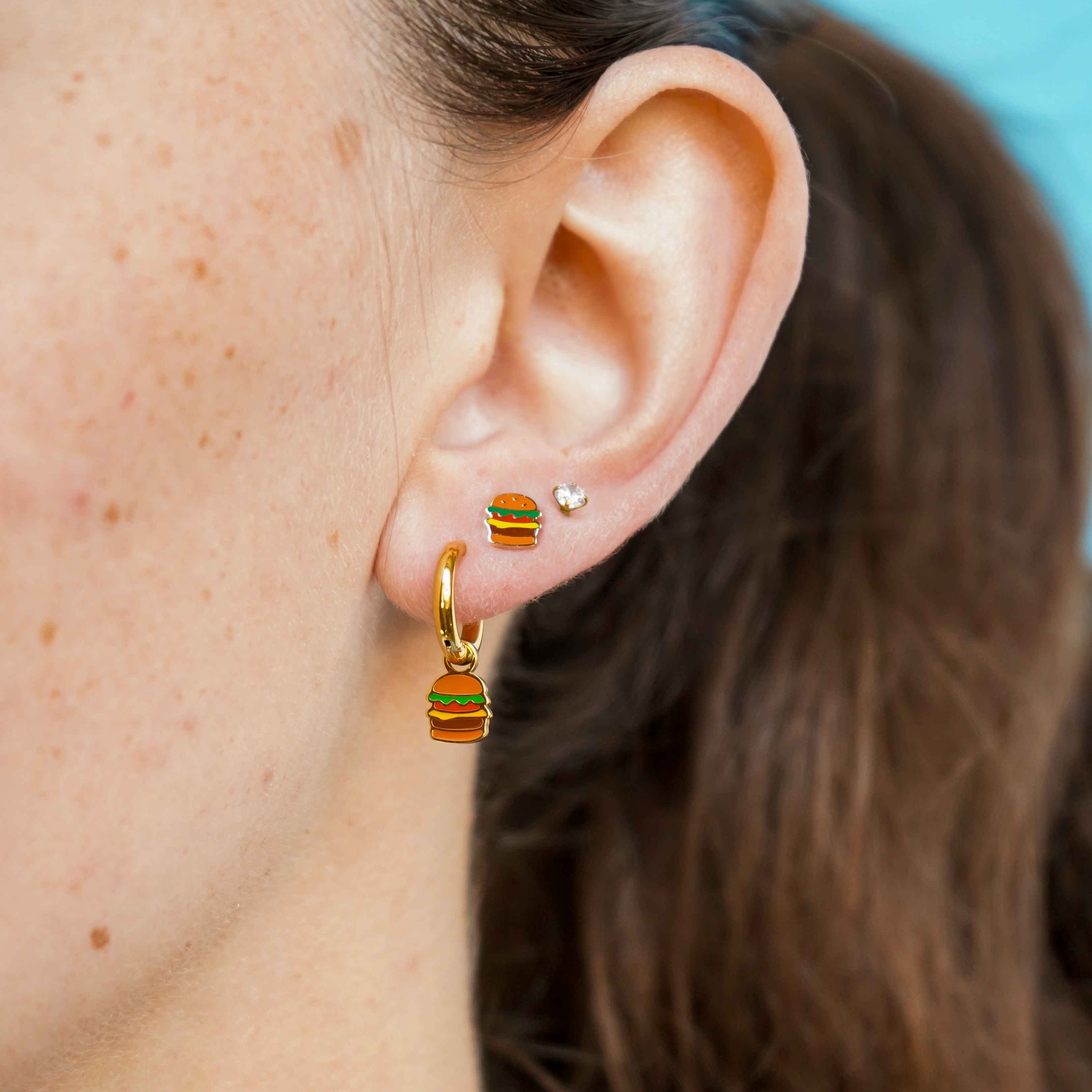 Close-up of an ear wearing a gold cheeseburger-shaped flat back stud earring and a gold hoop earring with a blue background