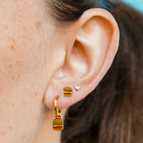 Close-up of an ear wearing a gold hoop with a cheeseburger-shaped charm on it with a blue background