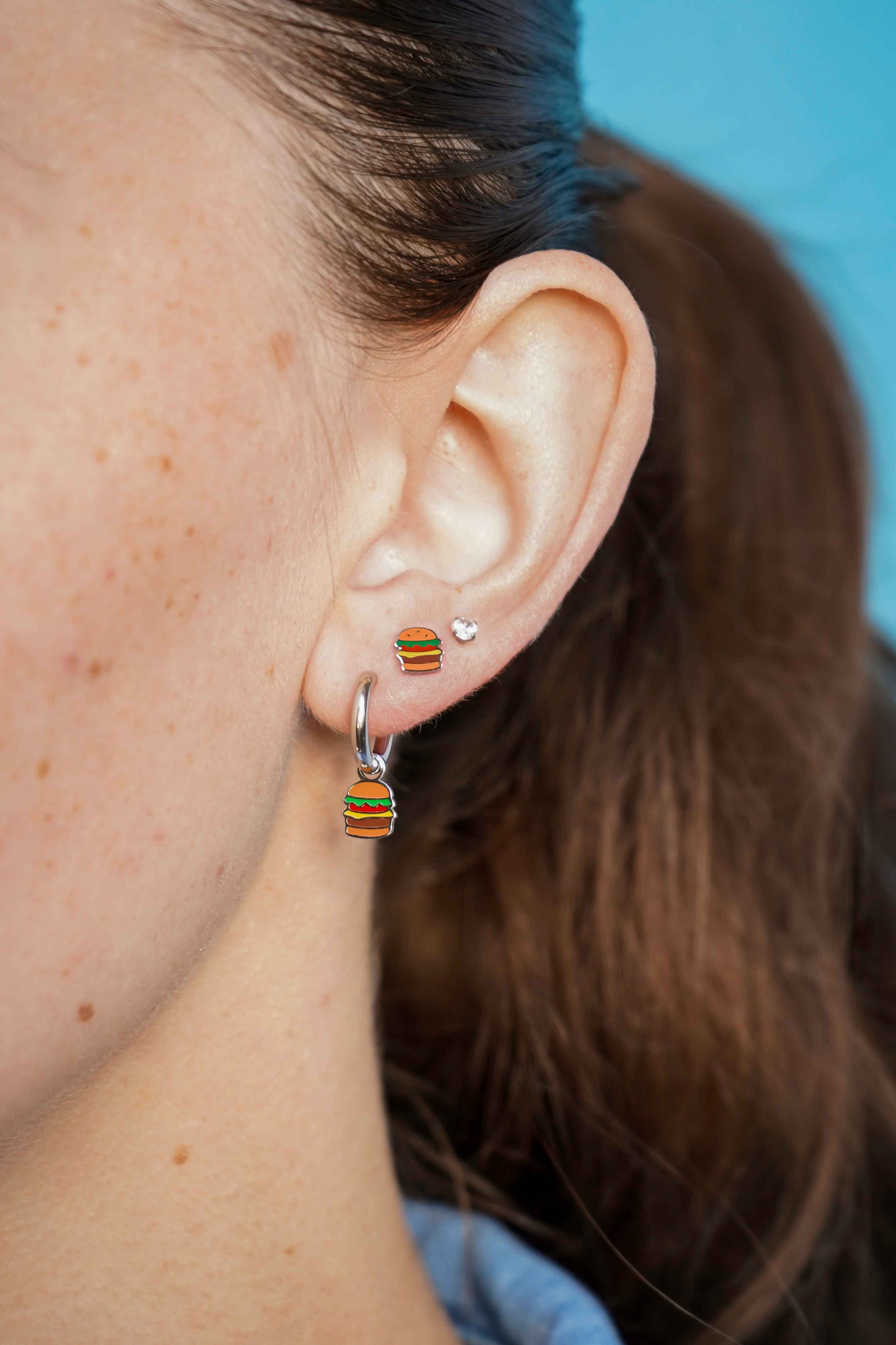 Close-up of an ear wearing a silver cheeseburger-shaped flat back stud earring and a silver hoop earring with a blue background