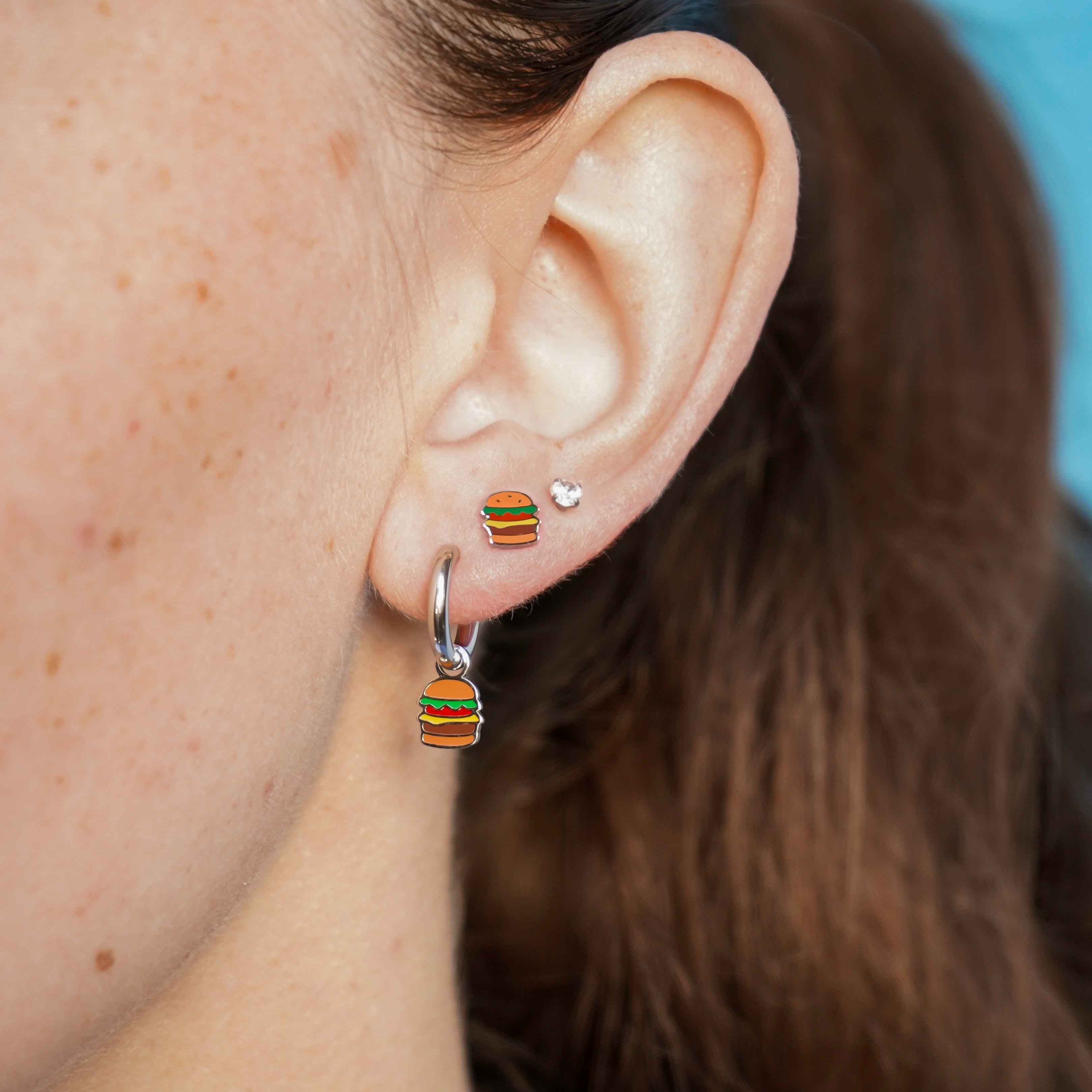 Close-up of an ear wearing a silver hoop with a cheeseburger-shaped charm on it with a blue background
