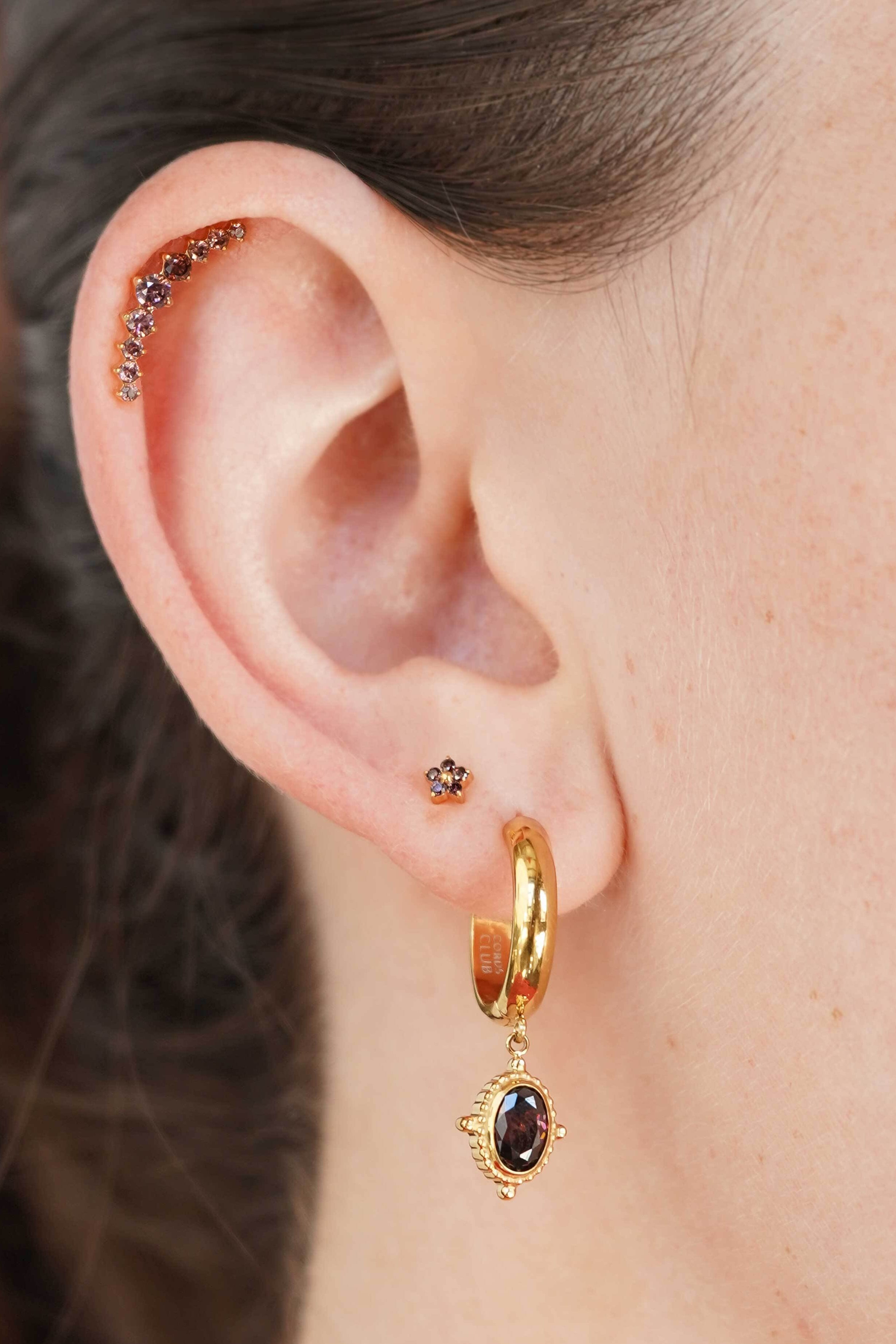 Close-up of a person's ear showcasing a multi-piece gold ear cuff with small dark gemstones, a small gold stud, and a gold huggie hoop with a dangling gold charm featuring a dark oval stone.