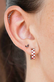 Close-up of a person's ear showcasing multiple piercings: a curved helix climber flat back stud with small brown stones, a small duo leaf stud, and a checkered hoop earring in silver and deep brown.
