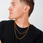 A man in a black t-shirt wearing two layered gold chain necklaces, shown in profile against a light gray background.