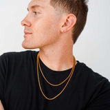 A man in a black t-shirt wearing two layered gold chain necklaces, shown in profile against a light gray background.