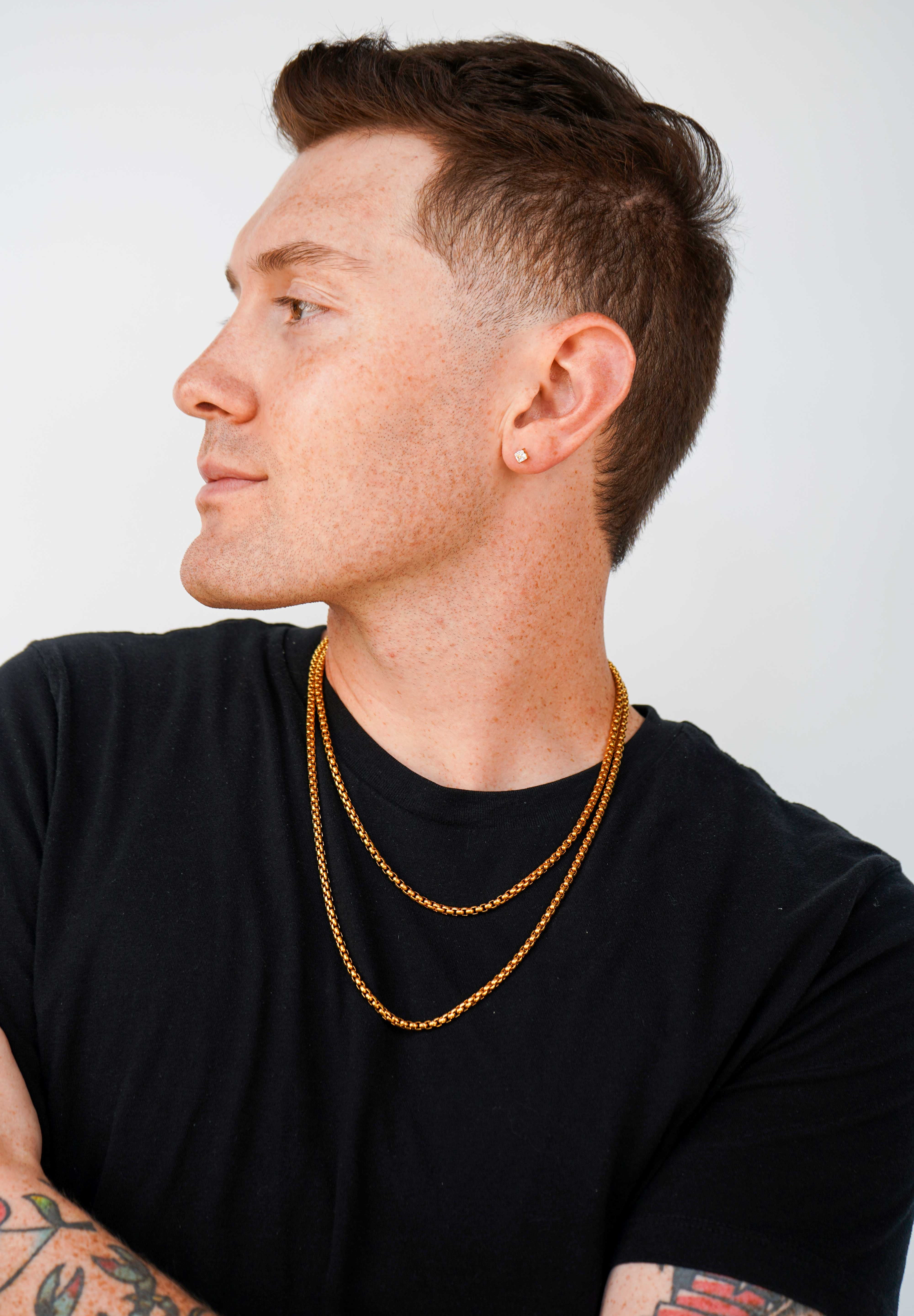 A man in a black t-shirt wearing two layered gold chain necklaces, shown in profile against a light gray background.