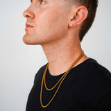 A man in profile wearing a layered gold chain necklace and a small gold stud earring, wearing a black t-shirt against a light gray background.