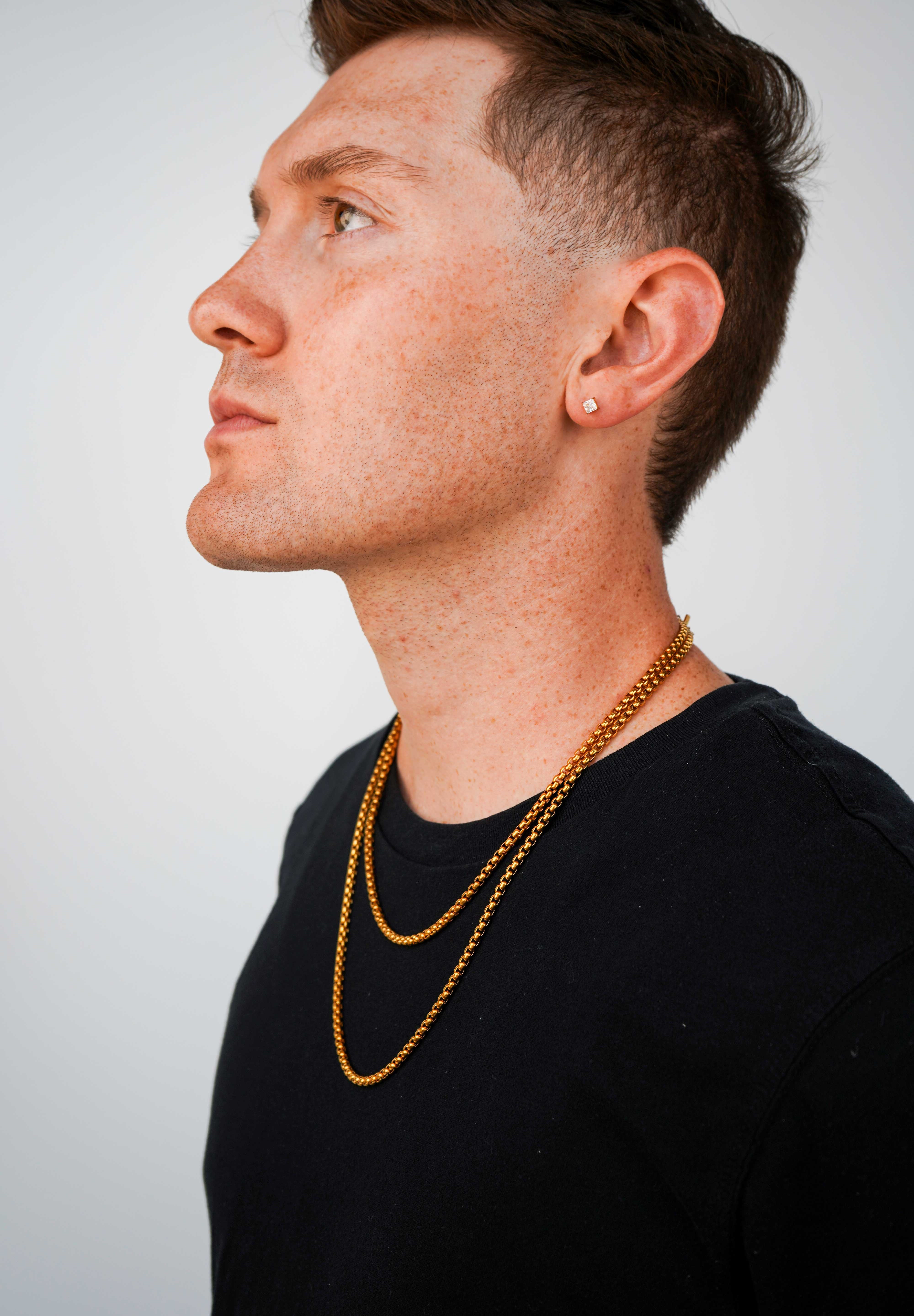A man in profile wearing a layered gold chain necklace and a small gold stud earring, wearing a black t-shirt against a light gray background.